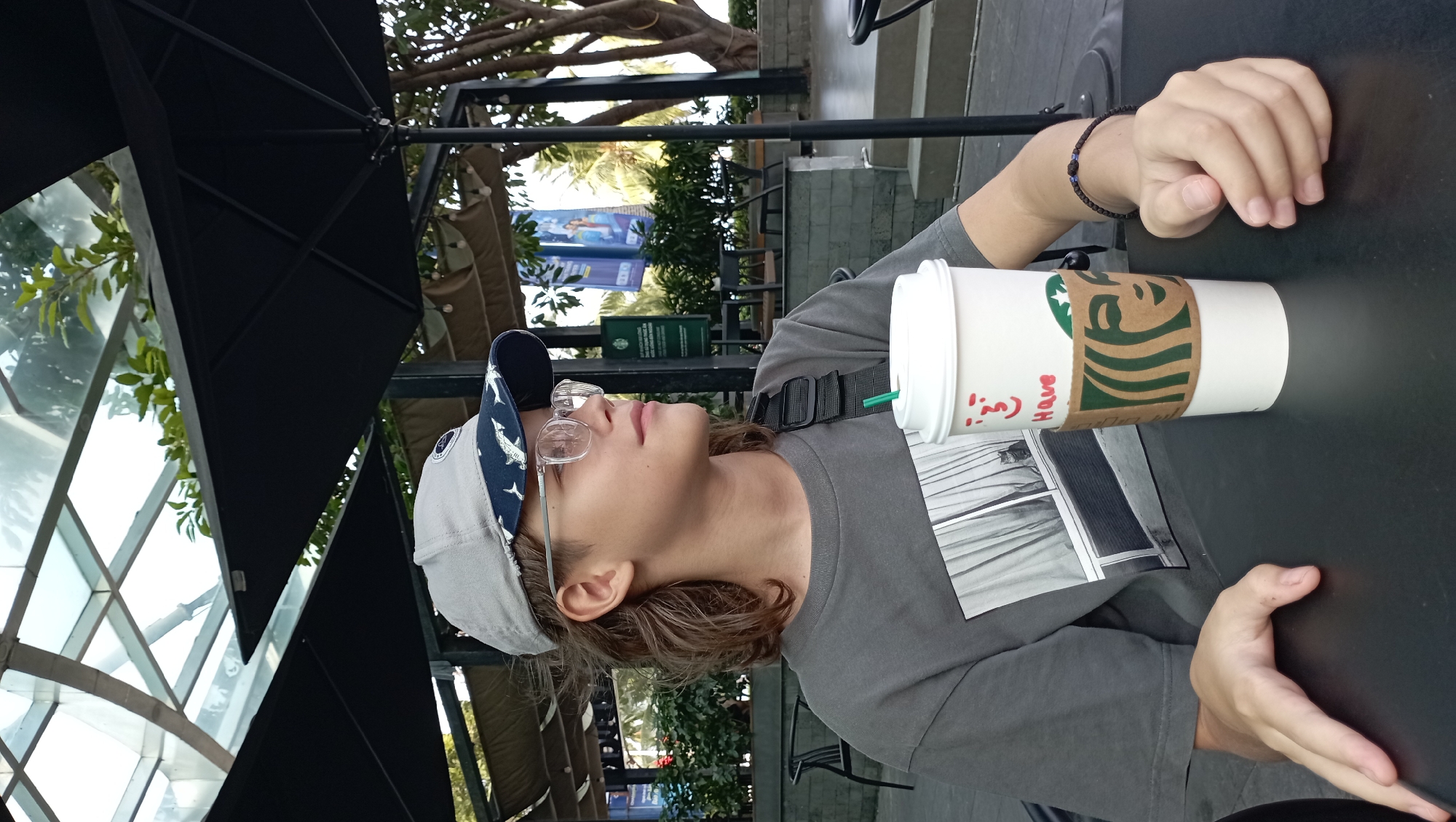 A person is sitting at a table outdoors at a Starbucks in Nha Trang Ward, Vietnam. The person is wearing a gray t-shirt, a baseball cap with a shark pattern on the brim, and glasses. They have a Starbucks cup on the table and are resting their hands on the table. The Starbucks is located outdoors under a canopy, and there are trees and foliage visible in the background. Banners are visible in the background.