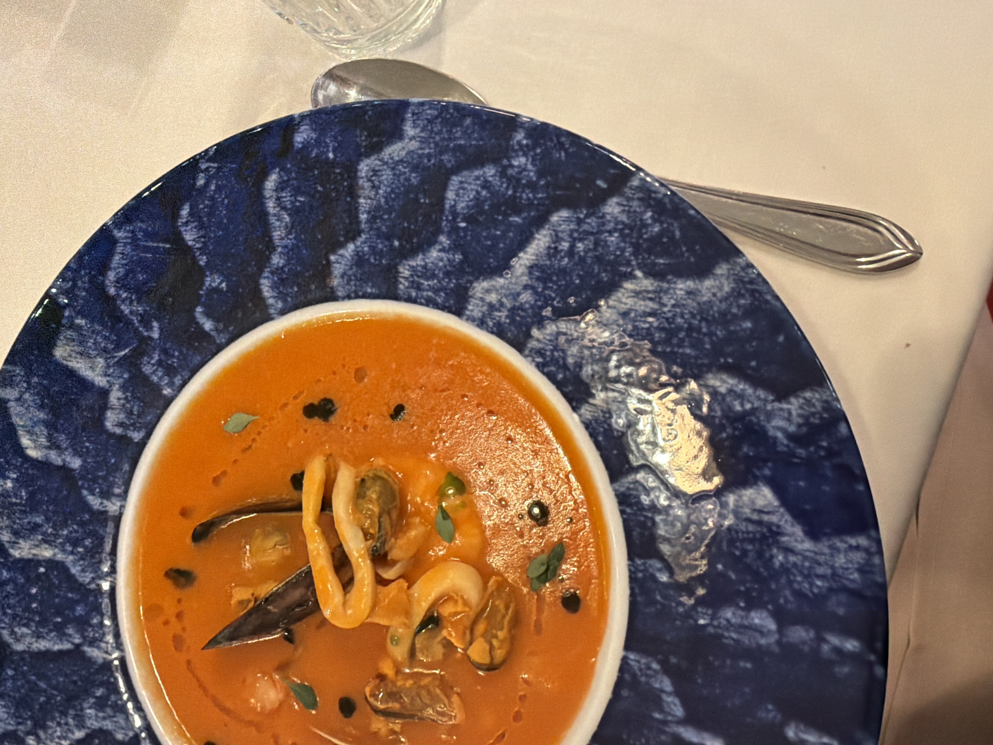 This close-up image captures a beautifully presented bowl of seafood soup, likely bouillabaisse or a similar dish, served in a restaurant in Yerevan, Armenia. The bright reddish-orange soup, appearing rich and creamy, fills a small white bowl. It contains a variety of visible seafood, including mussels (some in shells, some shelled) and white, curled squid rings, suggesting a fresh and substantial meal. The soup is garnished with small green herb leaves, possibly oregano or thyme, and tiny black dots, which could be pepper, black caviar, or another fine spice, adding visual interest and flavor.
The white soup bowl is nestled within a larger, decorative dark blue plate, which has a distinctive textured or wave-like pattern in varying shades of blue and black. This ornate plate rests on a crisp white tablecloth, suggesting an upscale dining environment. A silver-colored spoon lies partially visible on the tablecloth, to the right of the plate, glinting from the indoor lighting. Part of a clear glass can also be seen in the upper left corner. The lighting is soft, indicative of an indoor setting, likely for dinner or a formal lunch. There is no visible text in the image.