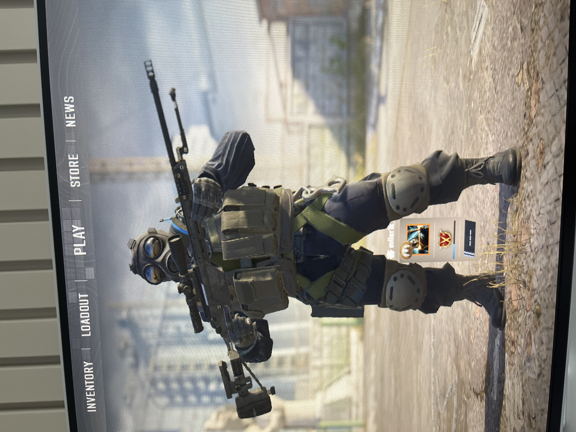 A digital screen shows a character in military gear against an urban backdrop. The character wears a gas mask, body armor with pouches, knee pads, and boots, and carries a rifle and a handheld device. On the left of the screen is a vertical menu with options like "Inventory", "Loadout", "Play", "Store", and "News." The background appears to be a dilapidated urban environment. Overlaid on the scene are some usernames, avatars and other GUI elements. The image does not contain the location in the city of Varna, Bulgaria.