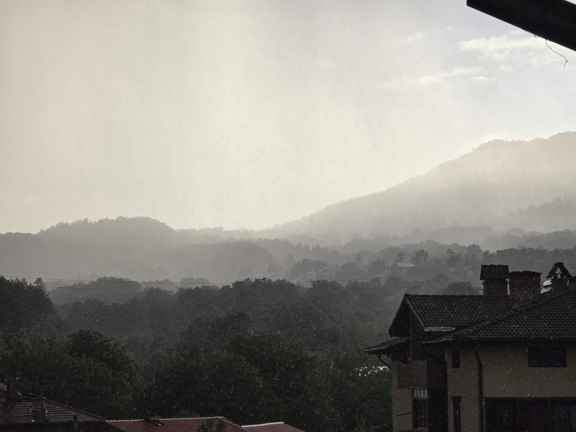 The image depicts a rainy day view in the city of Troyan, Bulgaria. In the foreground, sections of rooftops are visible, including a two-story building with a tiled roof and two brick chimneys. A dense forest stretches across the midground, with individual trees discernible. Beyond the forest, a mountain range is shrouded in mist and rain. The sky is overcast, contributing to the muted and greyish tones of the scene. The heavy rain obscures visibility.