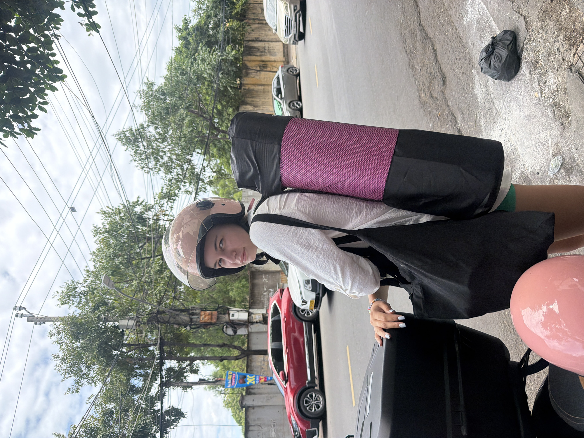 A young woman wearing a pink helmet, white long-sleeved shirt, and green shorts stands on a sidewalk next to a parked motorcycle in Nha Trang, Vietnam. She is carrying a rolled-up yoga mat strapped to her back and a black tote bag. In the background, there is a street with cars and trees. The sky is overcast.