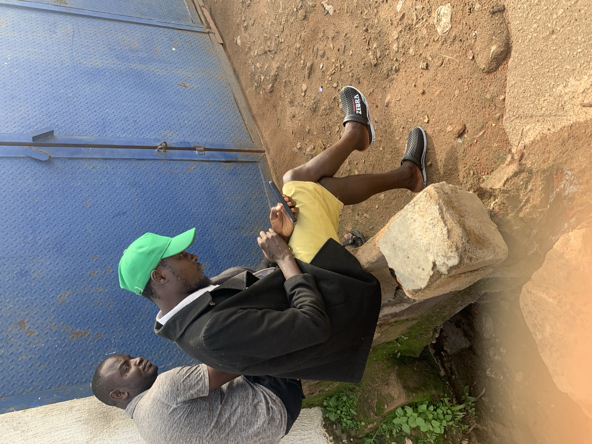 In Jos, Nigeria, two men are seen sitting near a blue metal gate with a textured diamond plate pattern. One man, on the left, wears a grey t-shirt and black shorts, looking towards the other man. The second man, in the foreground and facing right, is wearing a black jacket over a white shirt, yellow shorts, and black sandals with the brand "ZEBRA" visible. He is engrossed in his phone, which he holds in his right hand. The ground is composed of reddish-brown dirt and scattered stones. To the left, a rough concrete structure with some moss and greenery is visible, bordering a small stream or drainage channel. The scene appears to be outdoors, possibly on a street or in a more industrial area, given the gate and rough terrain.
