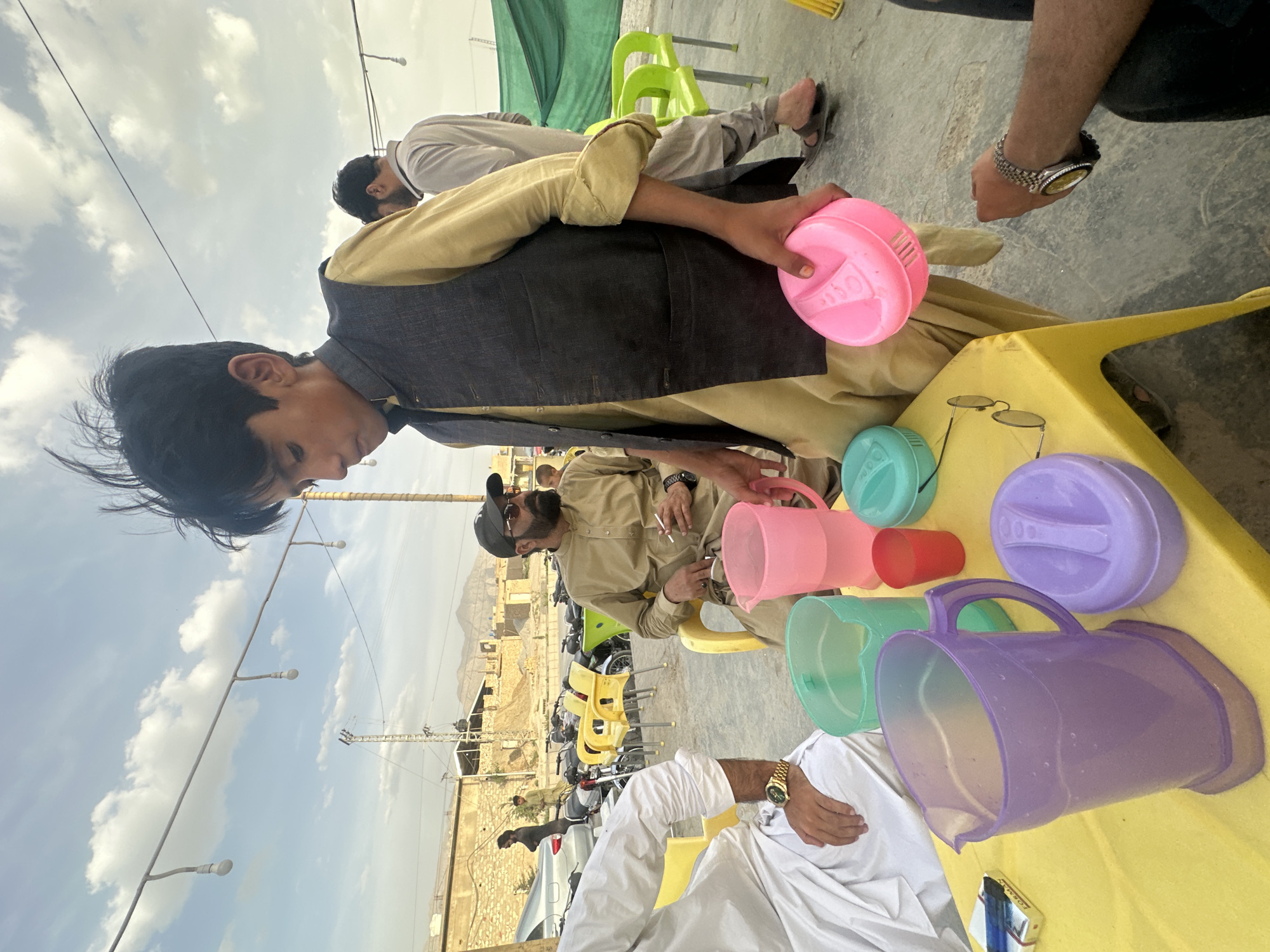 The image shows a group of men sitting outdoors at a gathering in Quetta City Tehsil, Pakistan. Several men are seated on yellow and green plastic chairs around a yellow table, which is covered with colorful plastic lids and jugs. The men are dressed in traditional Pakistani clothing. In the background, there is a landscape with mountains and buildings. The sky is partly cloudy.