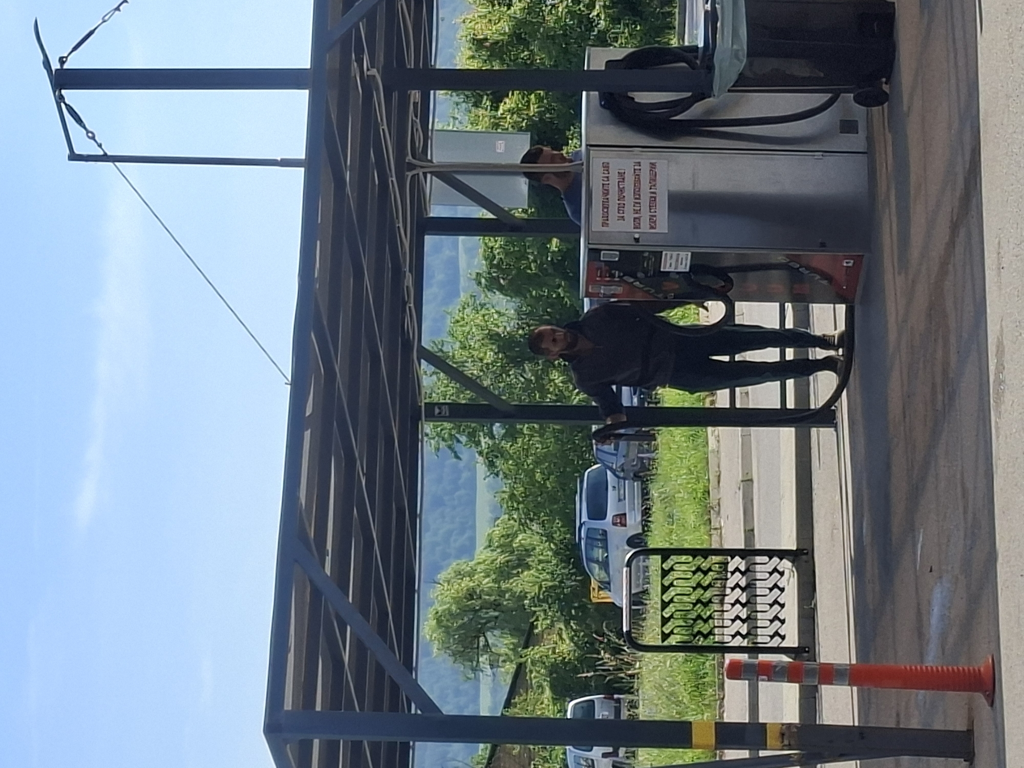 The image depicts a gas station in an unknown city. A man stands at a gas pump under the station's awning. In the background, vegetation and possibly a hill or mountain are visible. Several cars are parked near the station. Orange traffic barriers stand near the edge of the frame.