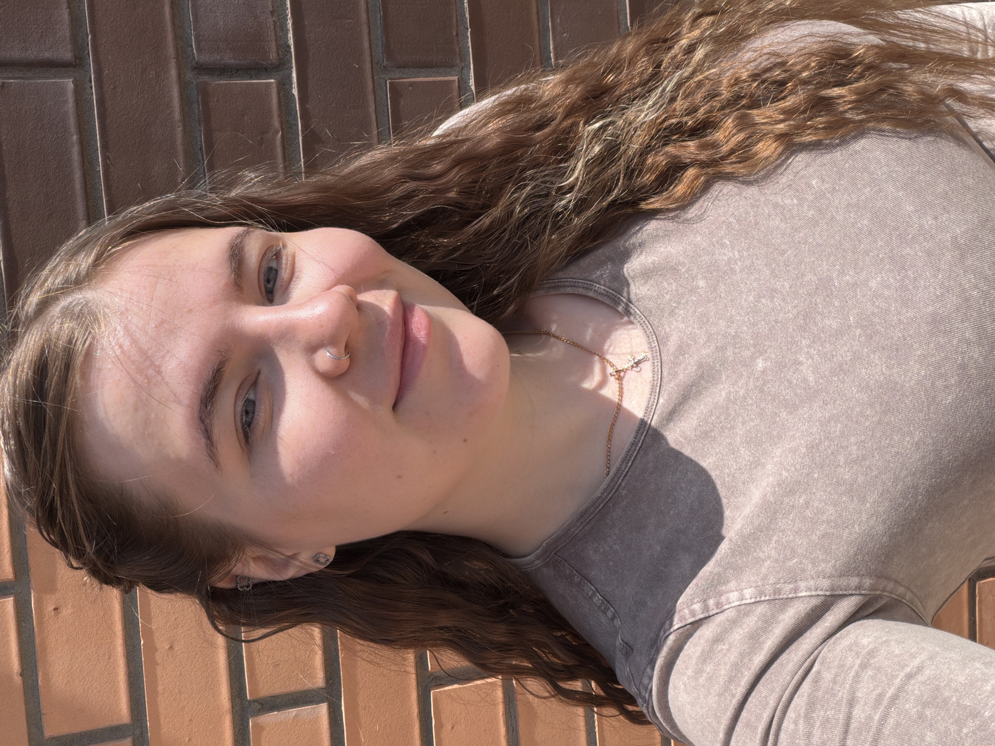 A young woman with long, curly brown hair poses for a photo. She wears a grey-brown, long-sleeved shirt and a gold necklace. A small gold nose ring is visible. She stands in front of a brick wall, presumably somewhere in the city of Chaikovsky, Russia.