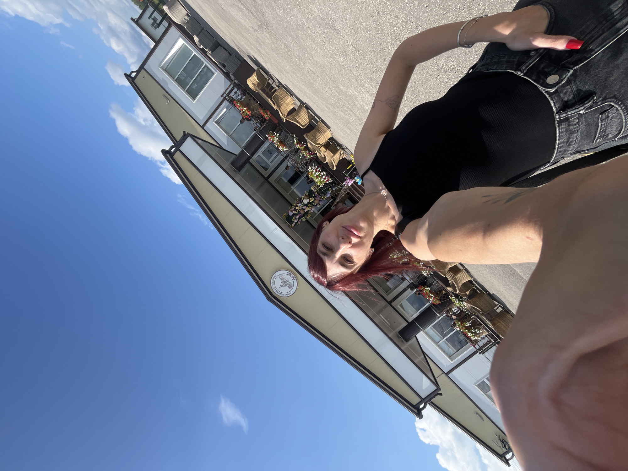 A woman with red hair and fair skin takes a selfie outdoors in front of a building. She is wearing a black tank top and black jeans. The building appears to be a commercial establishment, possibly a cafe or shop, with outdoor seating and potted plants. The sky is blue with scattered clouds. The image is slightly tilted. The location is identified as Akhlebaikha (Kovriginskii S/S), Russia.