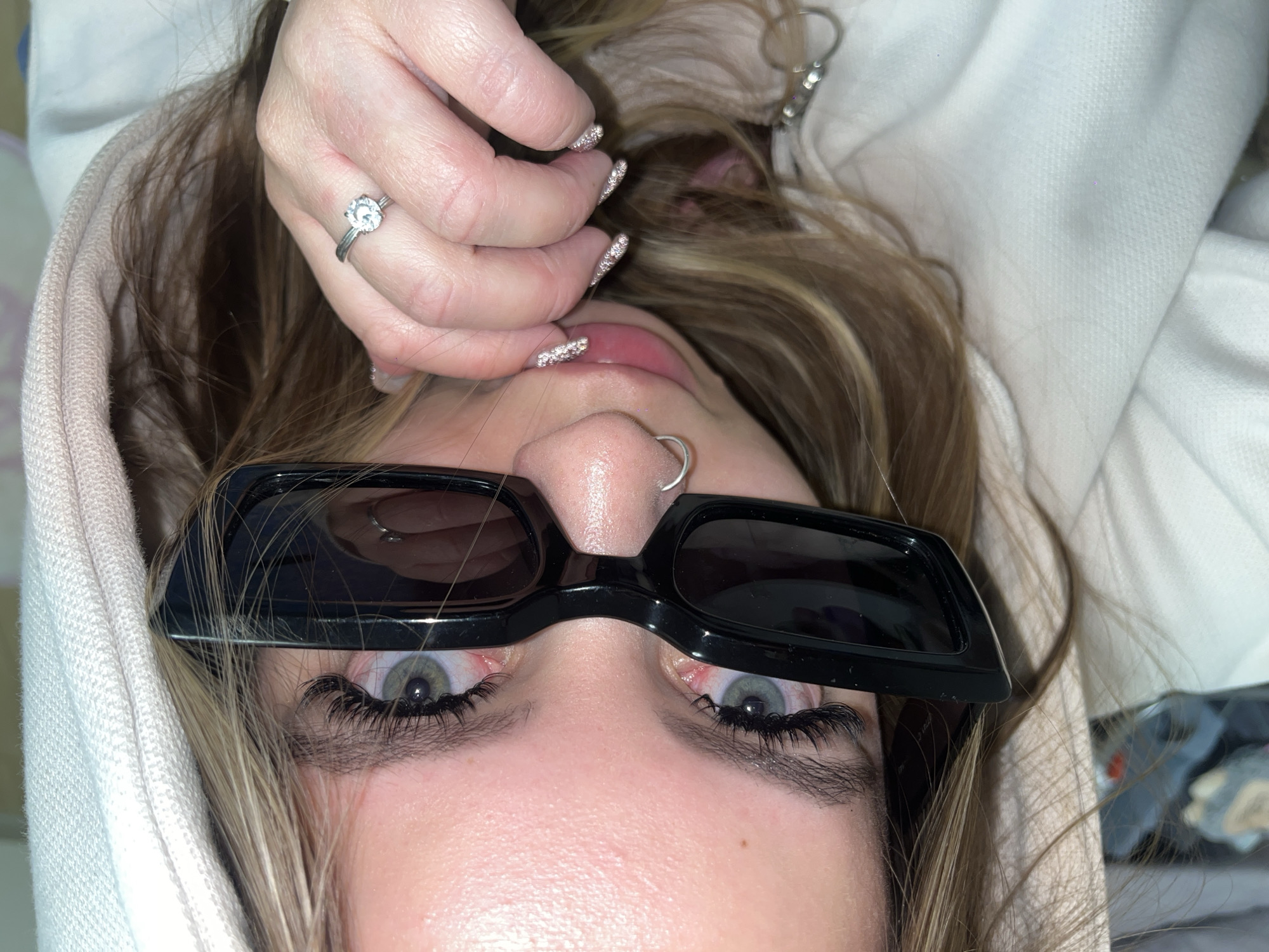 A close-up, high-angle photograph captures a person, likely female, lying on their back and looking directly upwards at the camera. The subject wears black, rectangular-framed sunglasses pushed down on the bridge of the nose, revealing light blue-green eyes with prominent, long dark eyelashes. A silver hoop nose ring is visible in the left nostril.
One hand is positioned near the mouth, showcasing long, glittery, light-colored fingernails and a silver-toned ring with a large, clear round stone on the ring finger. The lips are slightly parted, with a hint of the tongue showing. Long, highlighted blonde and light brown hair cascades around the face, partially framed by the hood of a light-colored, textured garment, possibly a white or pale pink hoodie.
The background offers limited detail, showing soft, blurry fabric or items on the right, suggesting a casual indoor setting. The image focuses tightly on the subject's face and upper body, indicating a posed shot. There is no visible text in the image. The location context is Krasnodar, Russia.