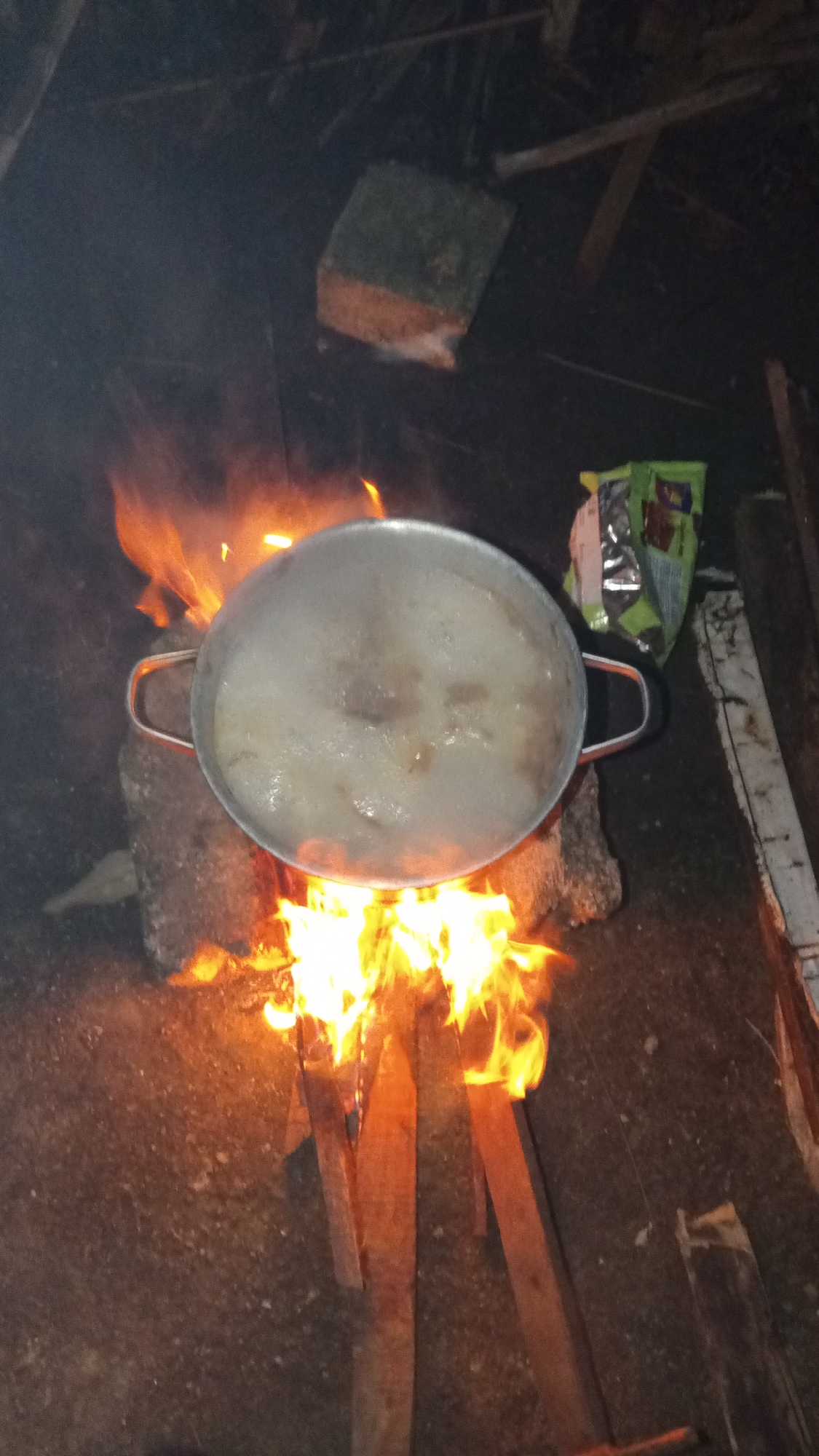 A silver pot of boiling liquid rests on top of stones above a small fire built of wood in Buketteukuh, Indonesia. A brick sits to the upper left of the pot. A green and silver bag lies next to a white board to the upper right of the pot. The ground is dirt. The background is dark.