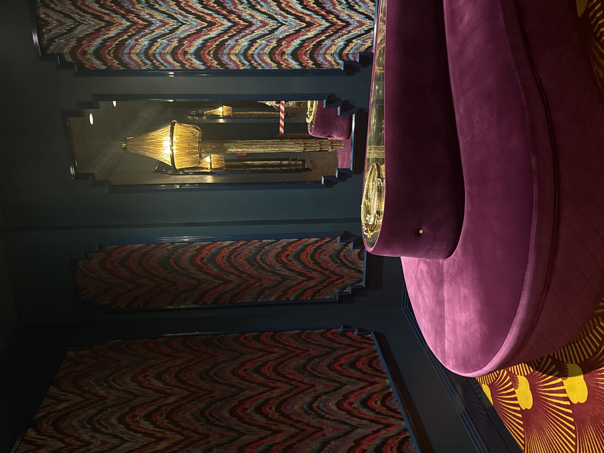 This media file captures an interior scene with bold, patterned decor and plush seating. The primary subjects are the decorative elements and the furniture. Several sections of wall or ceiling are adorned with a zig-zag pattern featuring a mix of red, blue, yellow, and black threads. One section of the wall exhibits a deep blue hue.
A prominent object is a large, curved piece of furniture upholstered in a vibrant, deep purple velvet. It appears to be part of a larger seating arrangement, possibly a sofa or banquette, with a sinuous, flowing design. A golden decorative trim with an ornate pattern is visible along the edge of the seating.
In the background, a mirror reflects light fixtures and more of the patterned walls. One light fixture is a large, glass, chandelier-style lamp with a golden glow. The reflection also shows what appears to be a section of another patterned wall or tapestry, and a vertical white and red striped object, possibly a barrier or marker. The overall impression is one of a luxurious and somewhat theatrical interior design.
There is no discernible activity or event taking place; the scene appears to be a still shot of the interior. The lighting suggests it is likely indoors, and the time of day is not evident. No specific location cues beyond the patterned walls and opulent furnishings are visible, and no emotions can be inferred from the static scene. No visible text is present in the image.