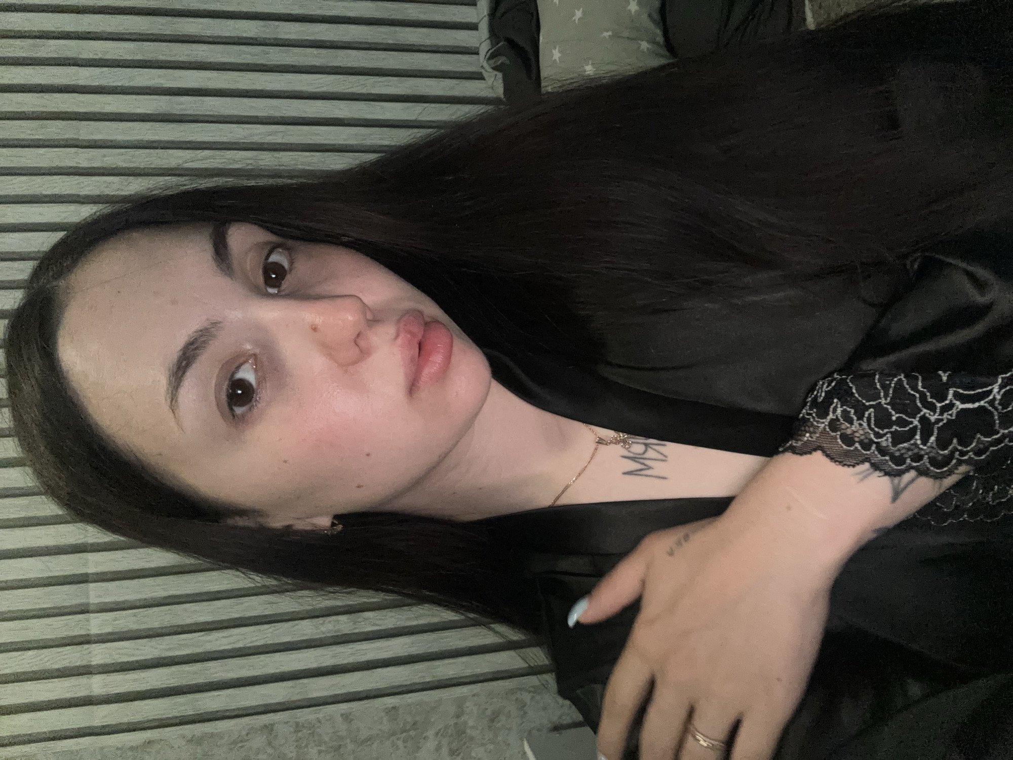 A young woman with long, dark hair and fair skin is shown in a selfie. She is wearing a black robe with lace trim. She has a necklace with a pendant and a tattoo on her chest. Her left arm is resting on her shoulder, and she is wearing a ring on her left hand. The background appears to be a room with a ribbed wall and a pillow with stars. The image does not provide any information about the location in the city of Марковский, Russia.