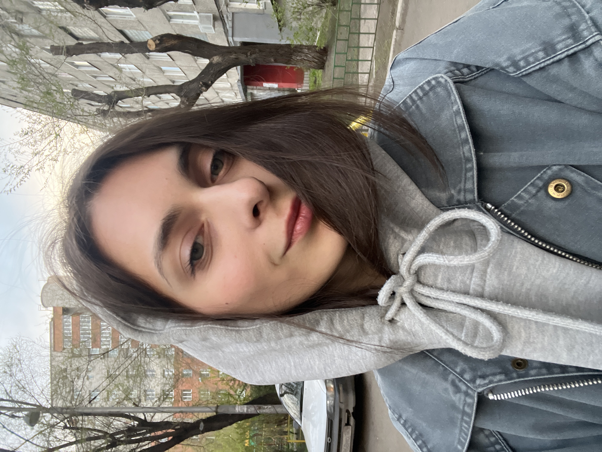 A woman with brown hair and a grey hoodie stands in an urban area. Behind her are several multi-story buildings, one with a red door and another with a lighter facade. Trees with bare branches are scattered around the buildings. The sky is overcast. A white car is partially visible in the lower right corner of the image. The woman is wearing a denim jacket over the hoodie. The location is Krasnoyarsk, Russia.