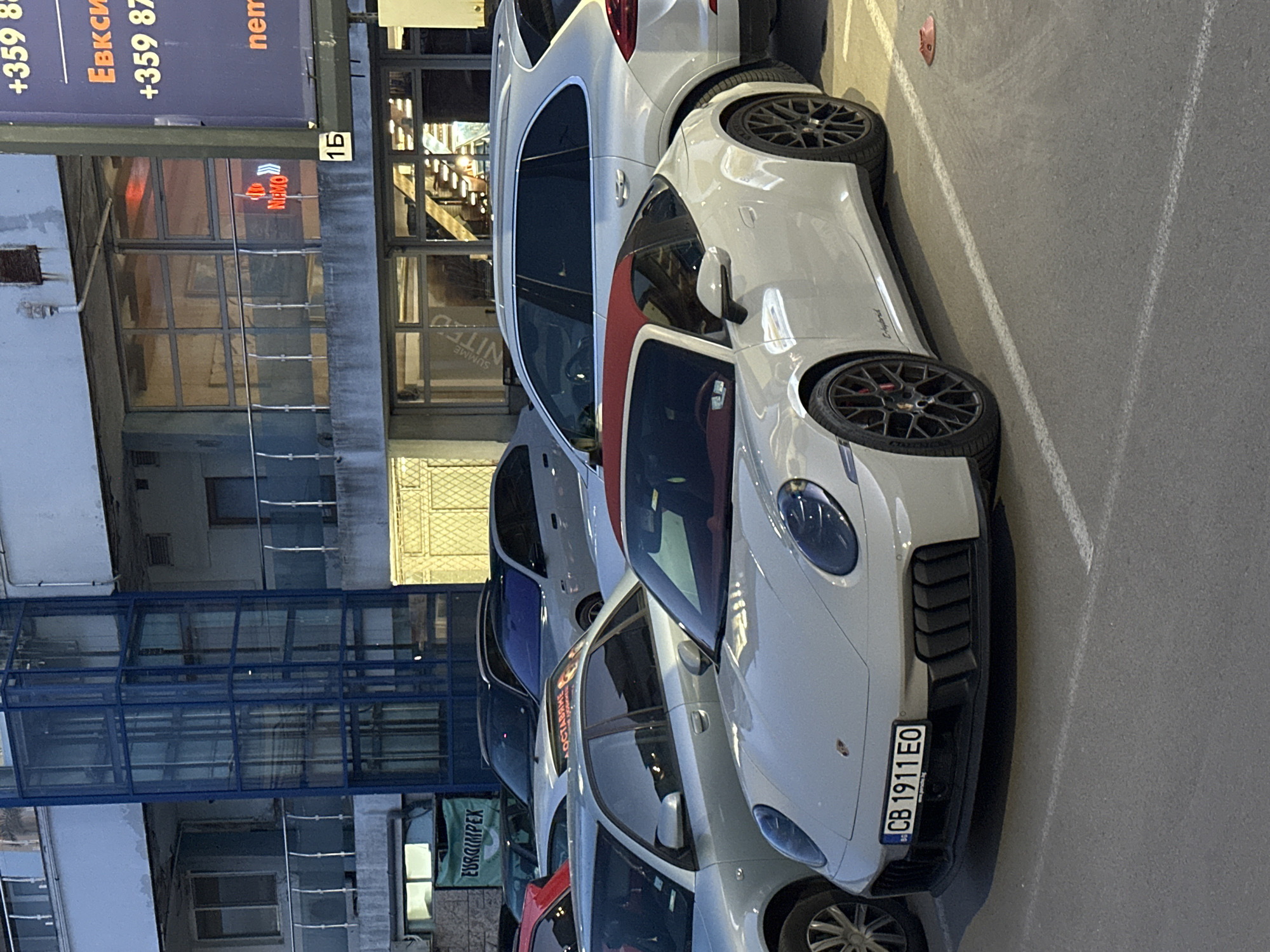 The image shows a street view, likely in the city of Varna, Bulgaria, with a row of parked cars along a curb. Prominently featured are several Porsche vehicles, including at least one Porsche 911 and a Porsche Cayenne E-Hybrid. The 911 appears to have a red interior and racing stripe. The license plate of the 911 is visible and reads "CB 1911 EO", indicating registration in Bulgaria.
The cars are parked next to a building with glazed windows. The building appears to have a restaurant or business called "NEMO" based on the sign. Another sign indicates the presence of the business "EURGIMPEX". A sign with phone numbers "+359 8" and "Евкси +359 87" is visible as well. A glass-enclosed walkway bridges over the street level, suggesting an elevated pedestrian connection.