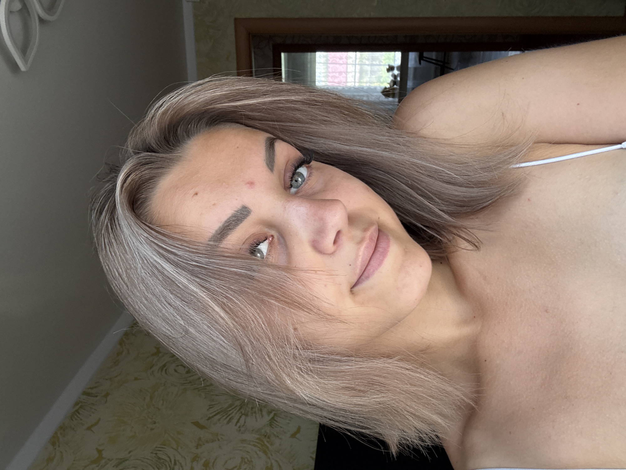 A young woman with shoulder-length, ash-blonde hair is posing for a selfie, angled slightly to her right. Her light blue eyes are looking upwards and to the left. She has light skin with some subtle freckles and a small blemish on her forehead. Her eyebrows are well-defined and dark. She is wearing a white top, the strap of which is visible on her shoulder.
The background suggests an indoor setting, likely a residential room. The wall behind her has a textured yellow and green wallpaper with a floral pattern. To the right, a doorway is visible, leading to another room. Through the doorway, a window with a lattice pattern and a glimpse of outdoor greenery can be seen. A piece of furniture, possibly a table with items on it, is also discernible. The lighting appears to be natural, coming from the window.
Based on the context provided, the location is the city of Krasnogorskoye, Russia. The scene is a personal moment captured in a home environment.
