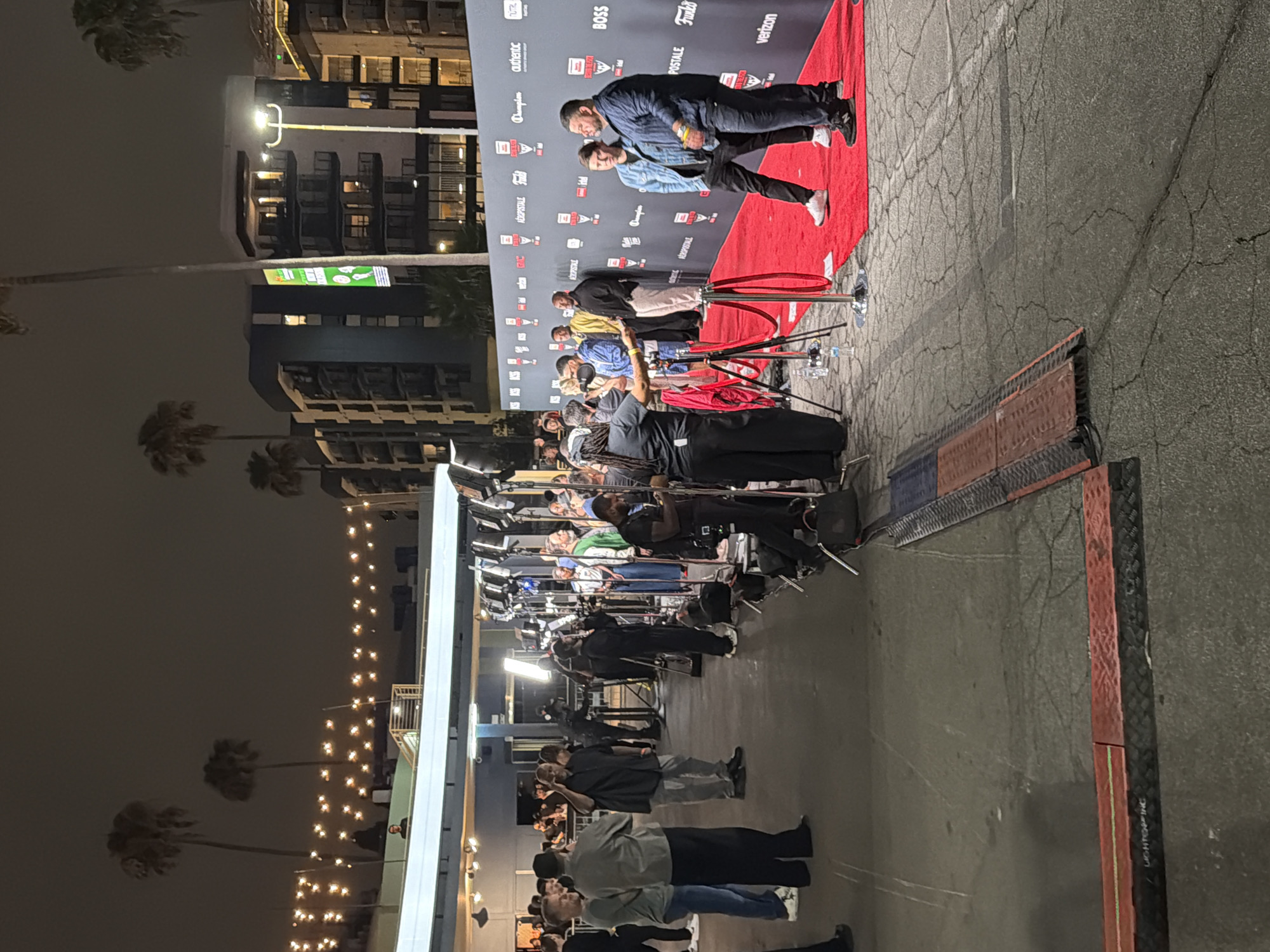The image depicts a red carpet event at night, likely in Los Angeles, California, United States of America, given the palm trees and urban architecture visible. The scene is set with a large red carpet leading up to a backdrop adorned with various logos, including "AEROPOSTALE," "Champion," "authentic," and "BOSS." A line of people, including media personnel with cameras and journalists, are positioned on the left side of the carpet, facing the backdrop. On the right side of the carpet, several individuals, presumably attendees or honorees, are posing for photographs and interacting with the media. The lighting is provided by numerous professional camera lights, illuminating the red carpet and the people. Palm trees and apartment buildings are visible in the background, suggesting an outdoor urban setting. The ground is paved asphalt.