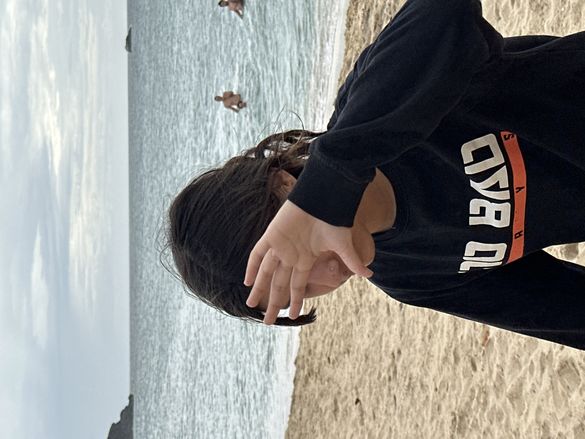 A young person is sitting on a sandy beach, with their hand covering their face. They are wearing a black long-sleeved shirt with white text that appears to say "TO BAD". The ocean is visible in the background, with several people swimming. The sky is overcast, and there is a rocky island in the distance. The image does not depict Altagracia, Venezuela; it is a generic beach scene.