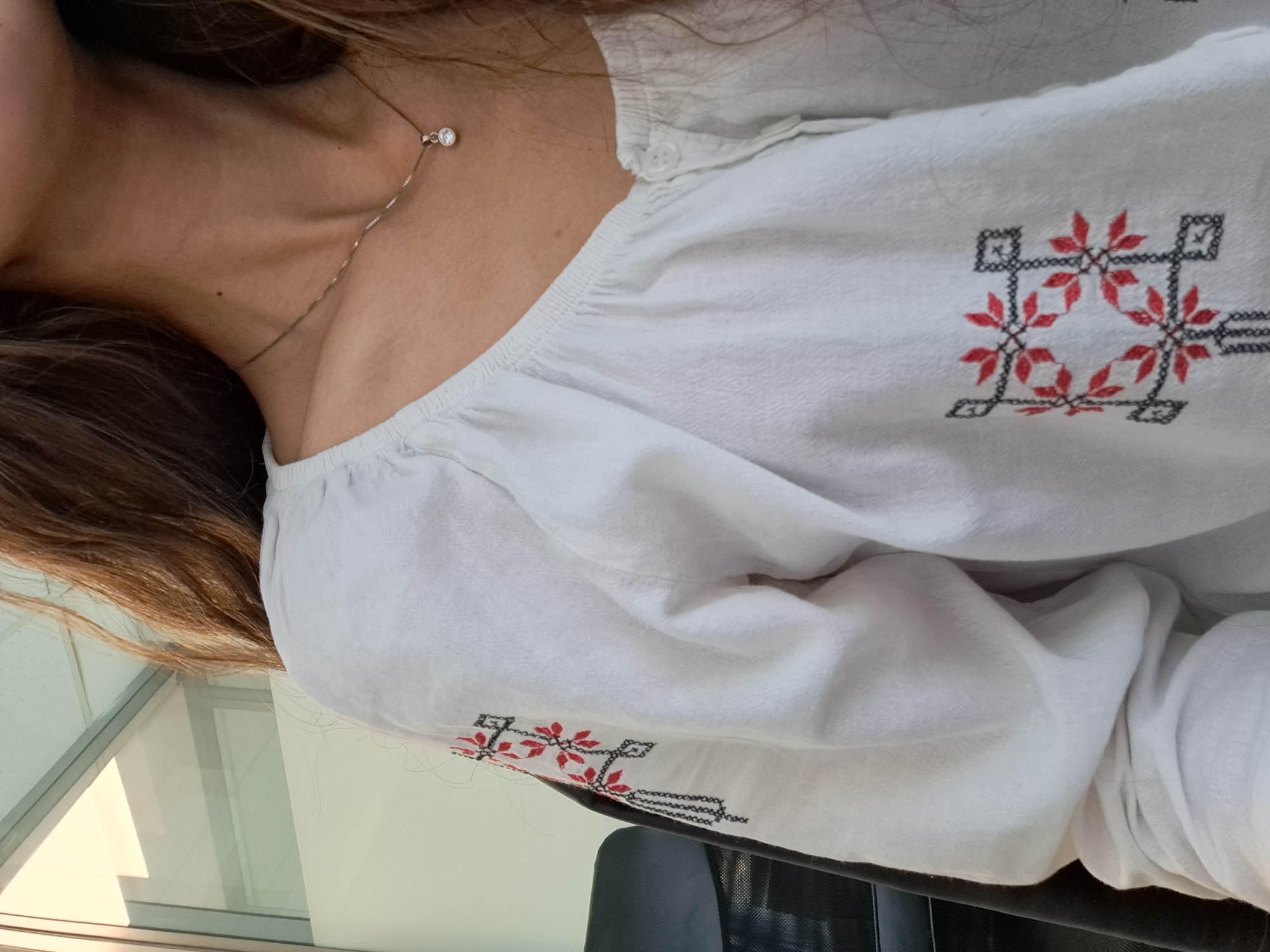 A person is wearing a white blouse with red and black embroidered details. The individual also has a thin necklace with a small pendant. The location appears to be indoors, possibly near a window. The city and specific location are unknown.