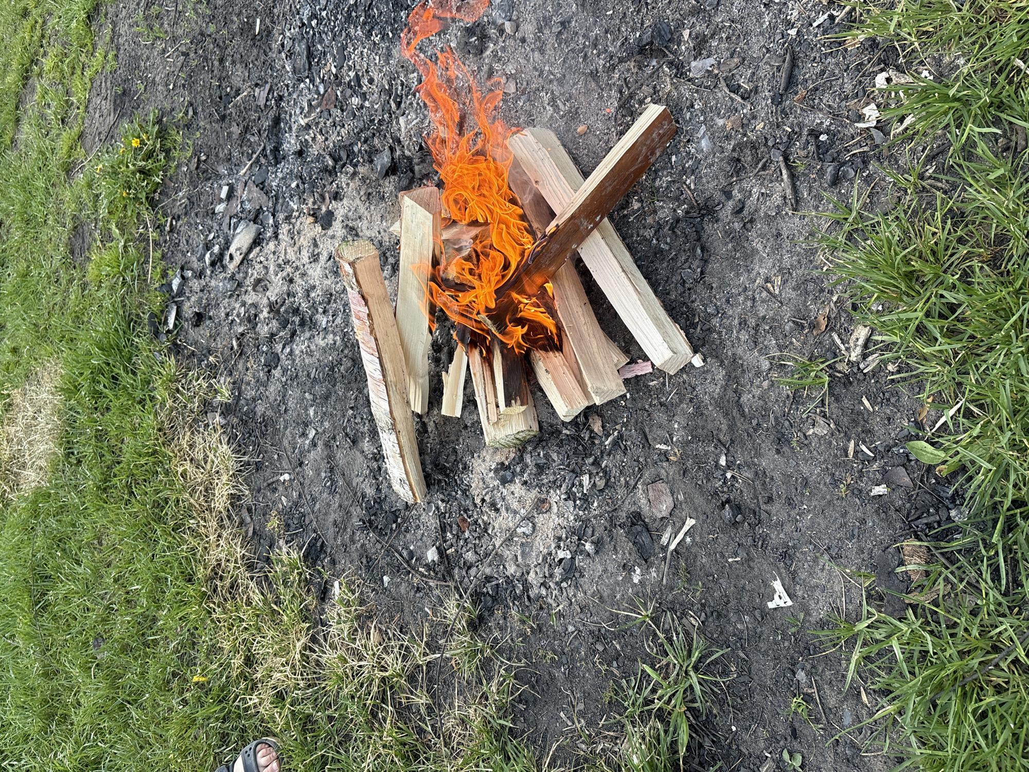 The image shows a small campfire in a clearing bordered by grass. The fire consists of stacked wooden logs with flames rising from the center. The ground around the fire is blackened with ash and charcoal remnants from previous fires. The edges of the clearing are defined by green grass, and a small portion of a sandal-clad foot is visible in the lower left corner of the frame. The precise location within Chaikovsky, Russia, cannot be determined from the image.