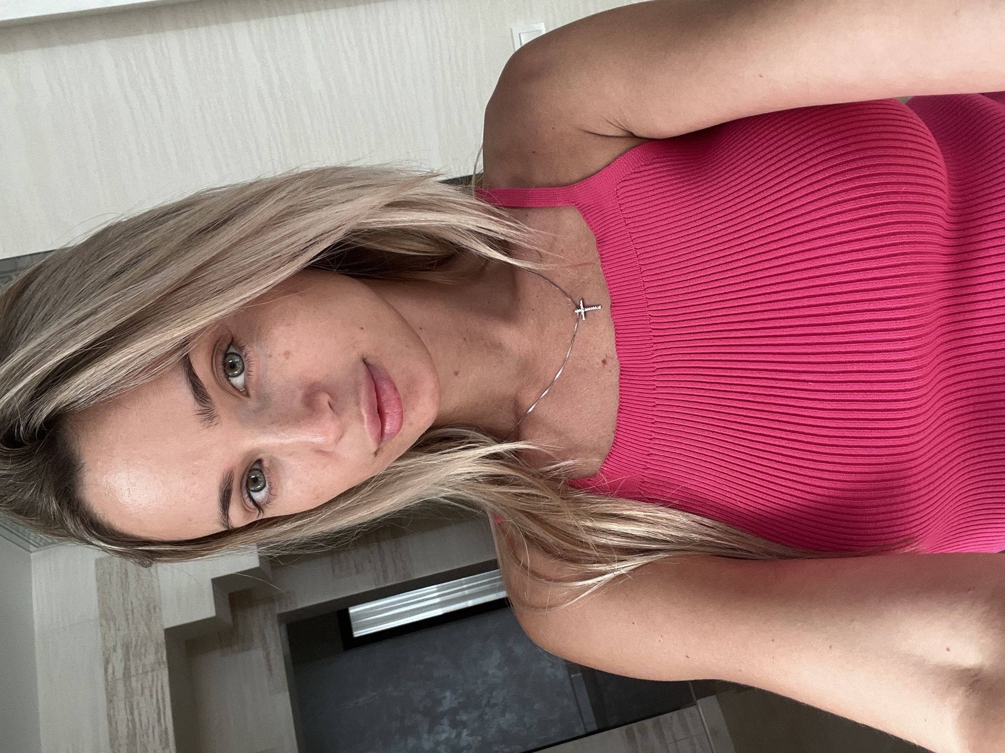 A woman with blonde hair and green eyes is taking a selfie in a room. She is wearing a pink ribbed tank top and a silver necklace with a cross pendant. The background of the image shows a wall with textured wallpaper and a dark doorway with vertical blinds. There is no indication that this image was taken in Fontanka, Ukraine.