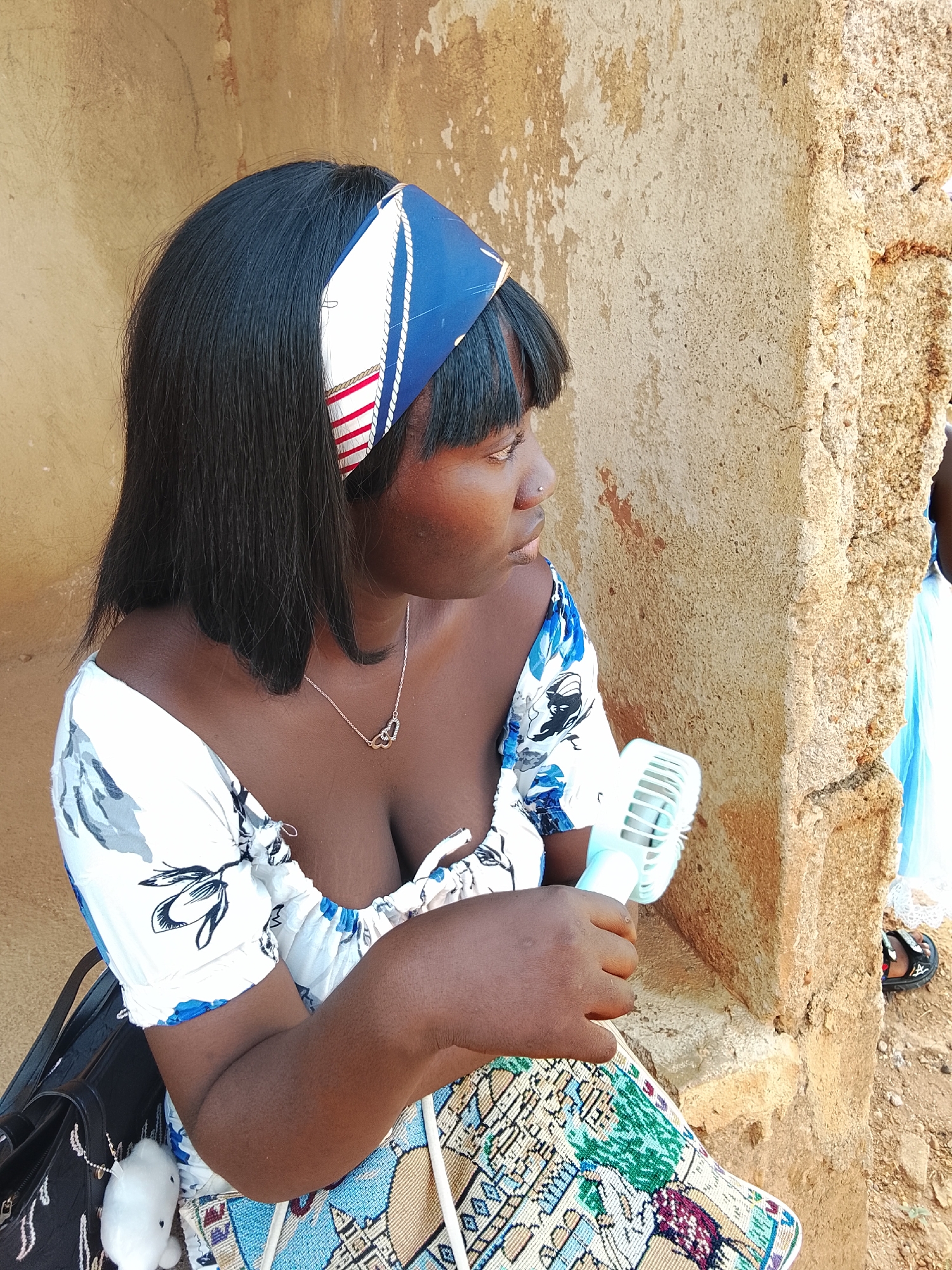 A woman is sitting outside a tan-colored building in Akwanga, Nigeria, wearing a white dress with blue floral patterns and a blue and white patterned headband. She has dark hair and is wearing a silver necklace. In her hands, she holds a small, light blue handheld fan. A black bag is next to her, with a white plush toy attached. The building has a weathered, textured surface. Another person is visible in the background wearing a blue dress and black sandal.
