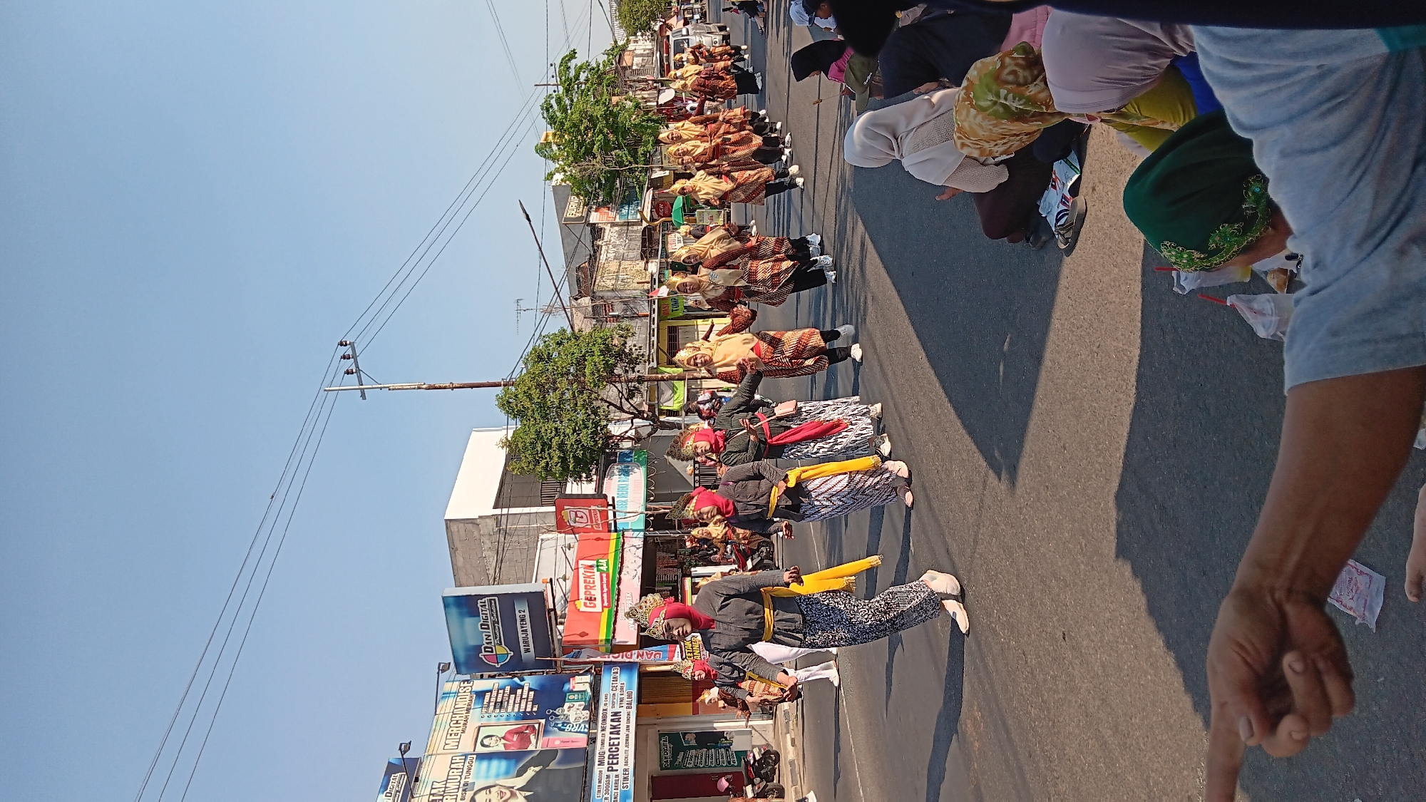 In the city of Tanjung Anom, Indonesia, a lively parade takes place on a sun-drenched street. A group of dancers, adorned in traditional attire consisting of grey tops, patterned sarongs, and colorful headscarves, moves in unison. They are holding hands and appear to be in mid-performance. Spectators, including women in hijabs, are seated on the side of the road, observing the procession. The background features buildings with various signs and awnings, indicating a commercial area. Power lines crisscross the clear blue sky above. The overall scene captures a vibrant cultural event in a bustling urban setting.