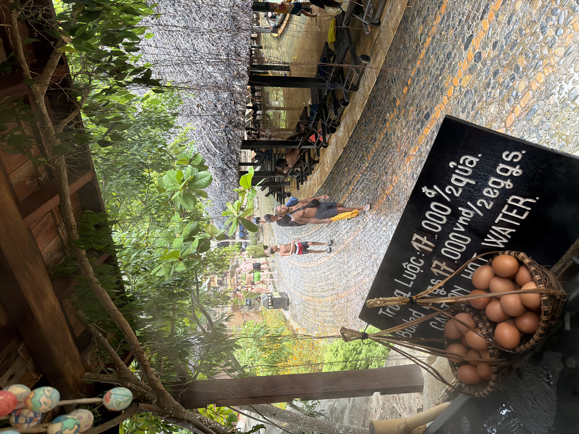 The image depicts a rainy outdoor scene at what appears to be a hot spring resort in Nha Trang, Vietnam. In the foreground, two woven baskets filled with brown eggs are suspended by ropes. To the right of the baskets, a black sign with white lettering lists prices in Vietnamese currency and English. The text reads "Trứng Luộc: 17.000 đ/2 quả, Boiled Eggs: 17.000 vnd/2 eggs, CÀNH NƯỚC NÓNG WATER".
The ground is paved with smooth, multi-colored stones that are wet from the rain, reflecting the surroundings. Lush green foliage hangs from a wooden overhead structure, and further back, thatched-roof shelters with lounge chairs are visible. Several people are present, some walking on the wet pathway and others resting under the shelters. The overall atmosphere suggests a tourist destination offering a unique experience, such as boiled eggs cooked in hot water.