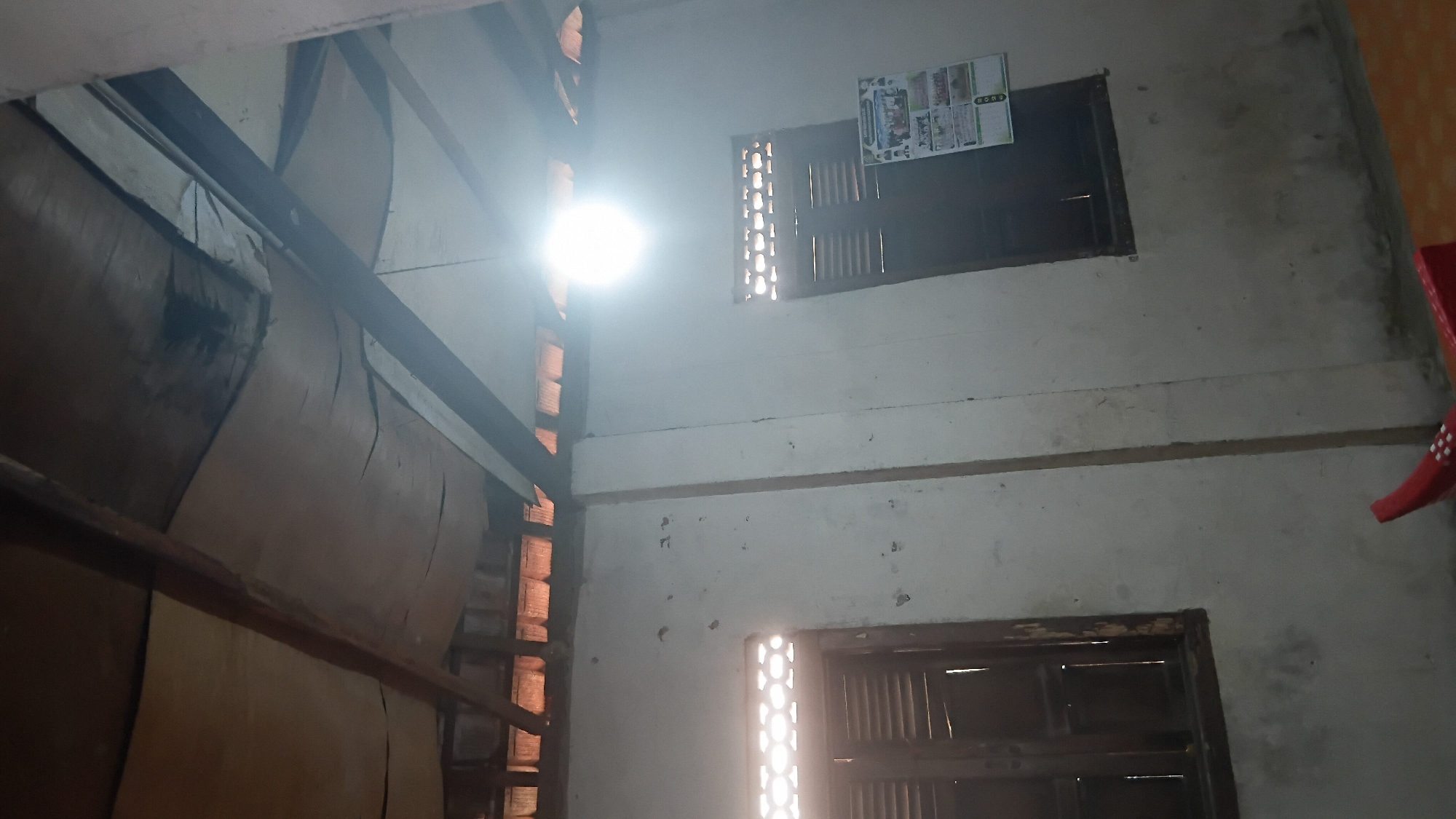 The image depicts the interior of a room with a rustic ceiling construction, possibly in Prabumulih, Indonesia. The ceiling consists of wooden beams supporting a light-colored material, which appears to be slightly damaged or discolored in places. Exposed roof tiles are visible above the beams.
On the walls, there are two windows with dark wooden shutters. The left window has a decorative grille pattern at the top, allowing light to pass through. The right window is similar, and above it, a calendar featuring images is affixed to the wall. The walls themselves are plastered and show some signs of wear or staining.
A bright light fixture is suspended from the ceiling, casting a strong glare in the upper right portion of the image. The floor appears to be a light-colored surface, possibly tiled or linoleum, with a hint of orange visible in the lower right corner. In the foreground, a red object with white markings is partially visible. The overall impression is of a simple, possibly rural or older dwelling.