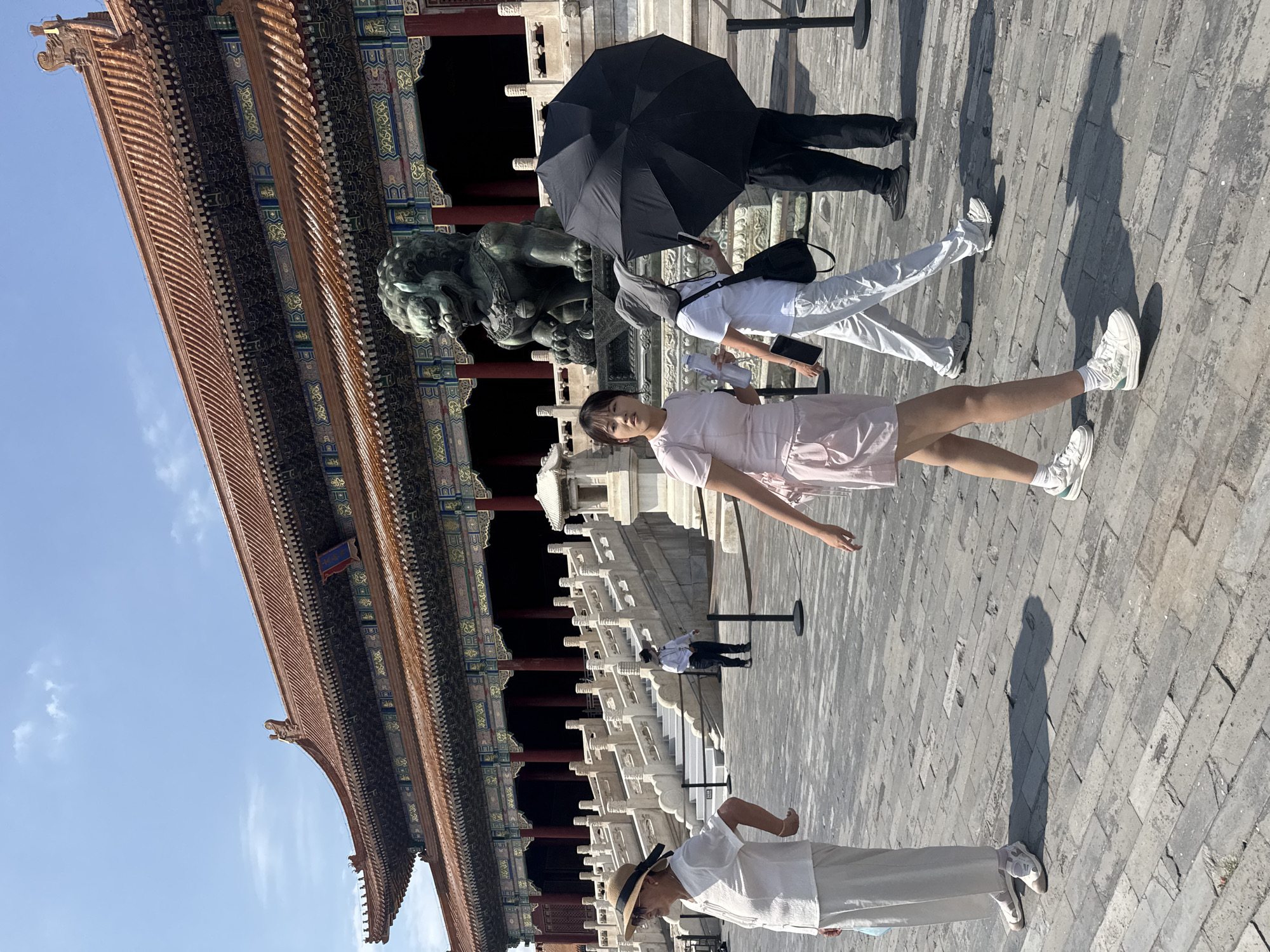 In Dongcheng District, China, a group of people are walking in an outdoor courtyard paved with stone bricks. In the background, a large, traditional Chinese building with a red tiled roof and ornate decorations is visible. A stone lion statue stands in front of the building. One woman, on the left, wears a straw hat, white shirt, and white pants. Another woman in the foreground wears a pink dress and white sneakers. To her right, another person holds a black umbrella, obscuring their face, while a fourth person in white pants and a hat walks alongside them. A fifth person in black pants and shoes is visible further to the right. The sky is blue with some clouds.