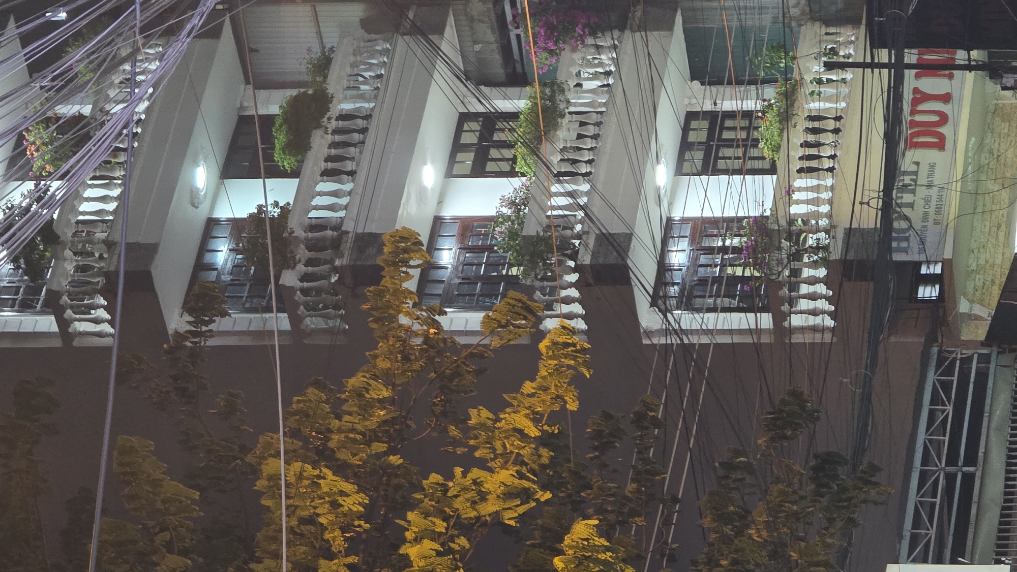 The image depicts an urban night scene in Bắc Nha Trang, Vietnam, characterized by buildings, illuminated trees, and a dense network of overhead wires.
The primary subjects are multi-story buildings, likely residential or hotels, featuring light-colored facades. Many of these buildings include balconies with distinctive white, decorative balustrades. Potted plants, including green shrubs and flowering varieties with pink and purple blossoms, are visible on the balconies, adding greenery to the architecture. The buildings feature numerous windows, some with dark brown frames and grid patterns, with interior lights casting a warm glow.
In the foreground, a cluster of trees with dense, broad leaves stands prominently. These trees are brightly illuminated by artificial light, giving their foliage a vivid golden-yellow hue against the dark, nighttime sky.
Above and around the buildings and trees, a thick tangle of black and grey electrical or communication wires crisscrosses the frame, a common feature of urban infrastructure in many Southeast Asian cities.
A significant detail is a large, vertically oriented sign on the side of one building, partially obscured by the wires. It clearly reads "HOTEL DUY M" (likely "HOTEL DUY MINH"). Below this, smaller Vietnamese text identifies the address: "K 90 NGUYỄN ĐÌNH CHIỂU - NHA TRANG" and provides a phone number: "ĐT: 0583 544 114." These details confirm the location as Nha Trang, Vietnam.
The scene captures the still, bustling atmosphere of a city at night, with artificial lighting highlighting both architectural elements and natural features under a clear, dark sky. There are no visible people or specific activities taking place.
