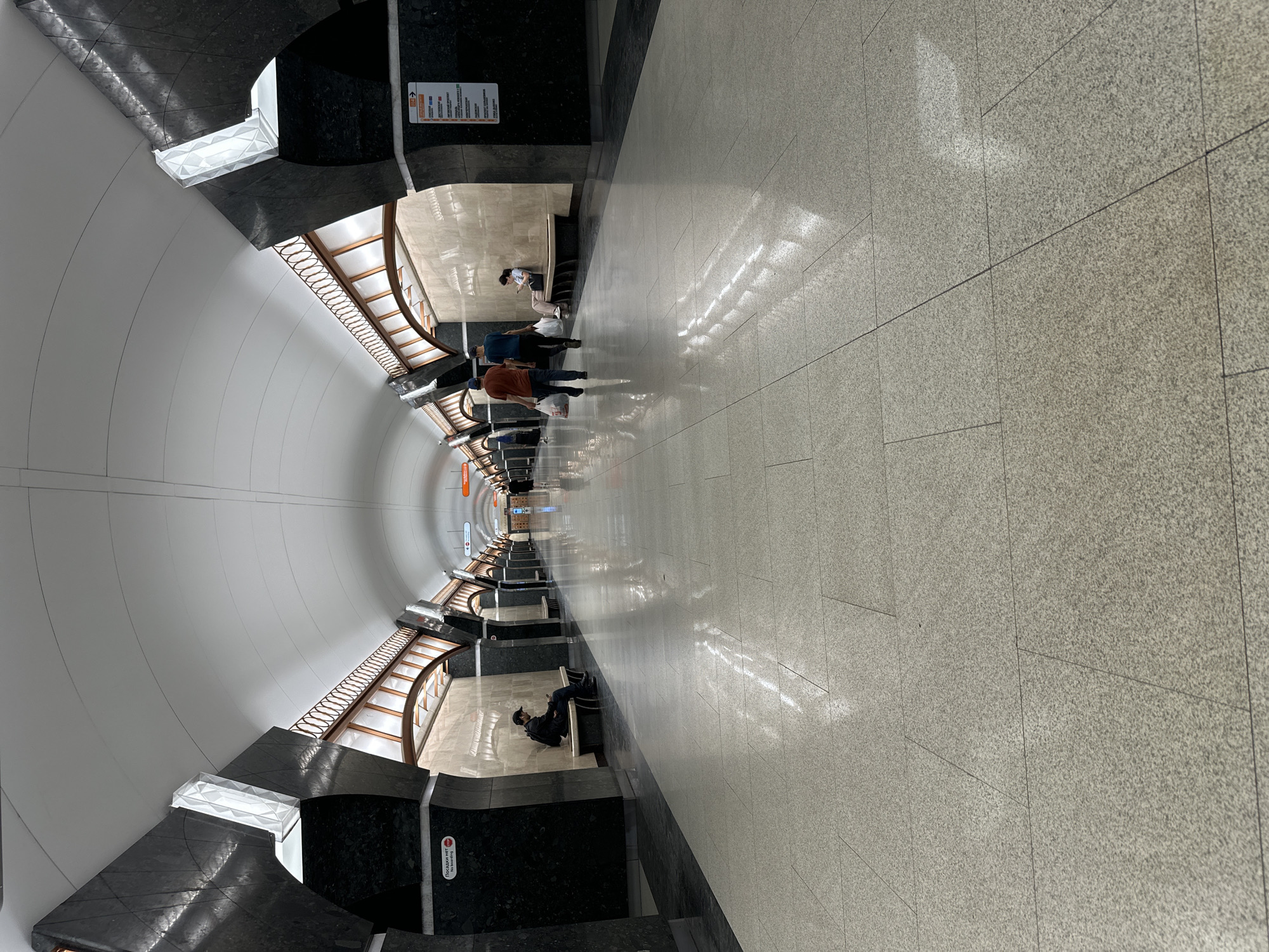 The image shows a view down a long, arched subway station platform in Saint Petersburg, Russia. The station features a high, white, arched ceiling with decorative lighting fixtures integrated into the architecture. The platform is paved with large, light-colored granite tiles that reflect the ambient light, creating a bright and spacious atmosphere. Dark, polished stone is used for the columns and the lower sections of the walls, providing a stark contrast to the lighter elements.
Several people are present in the station. On the left side of the platform, a man wearing a cap and dark jacket is seated on a bench, looking at his phone. Further down the platform, two men are walking towards the viewer, each carrying shopping bags. One is wearing an orange t-shirt and the other a blue t-shirt. A woman is seated on a bench to the right, also looking at her phone. Other indistinct figures can be seen further in the distance, suggesting ongoing passenger activity. Benches are spaced along the platform walls. Signage, some appearing to be in Russian, is visible on the walls, including directional signs and information boards. The overall impression is of a well-maintained and modern underground transit environment.