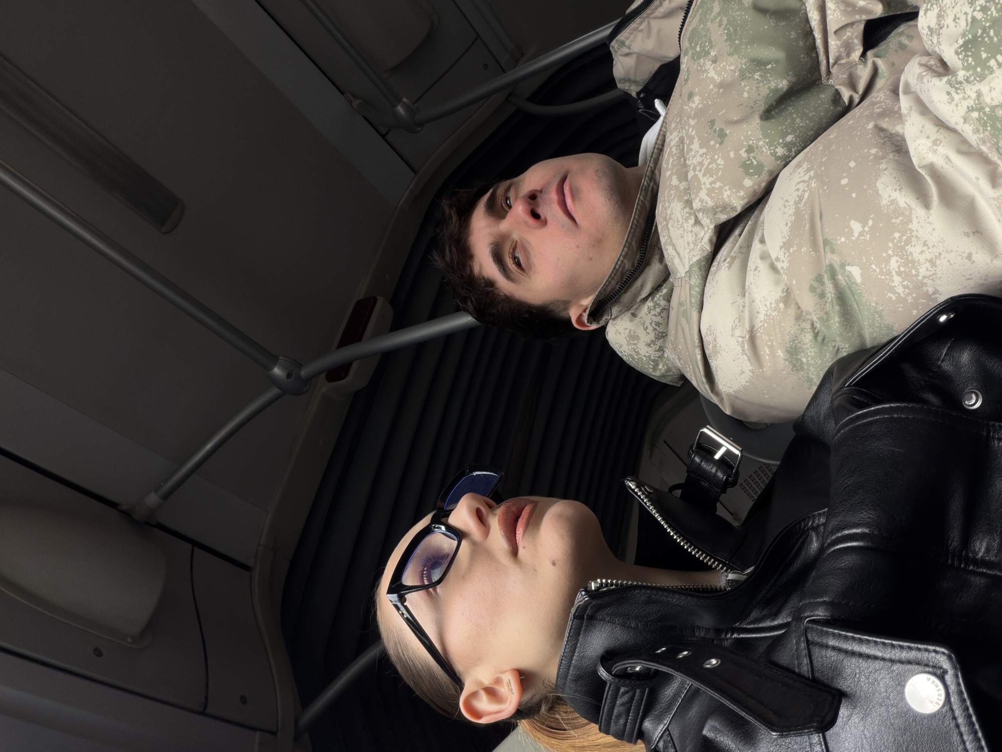 Two people are in what appears to be an airplane cabin or a similar enclosed space with overhead compartments and fixtures. The space seems cramped. A man wearing a camouflage-patterned puffer jacket is visible towards the upper center of the image. Below him is a woman wearing black-rimmed glasses and a black leather jacket. The camera angle is looking upwards at the two individuals. The interior of the aircraft or vehicle is a neutral color, potentially gray or white. The location of the photograph, Sofia, Bulgaria, is unknown.
