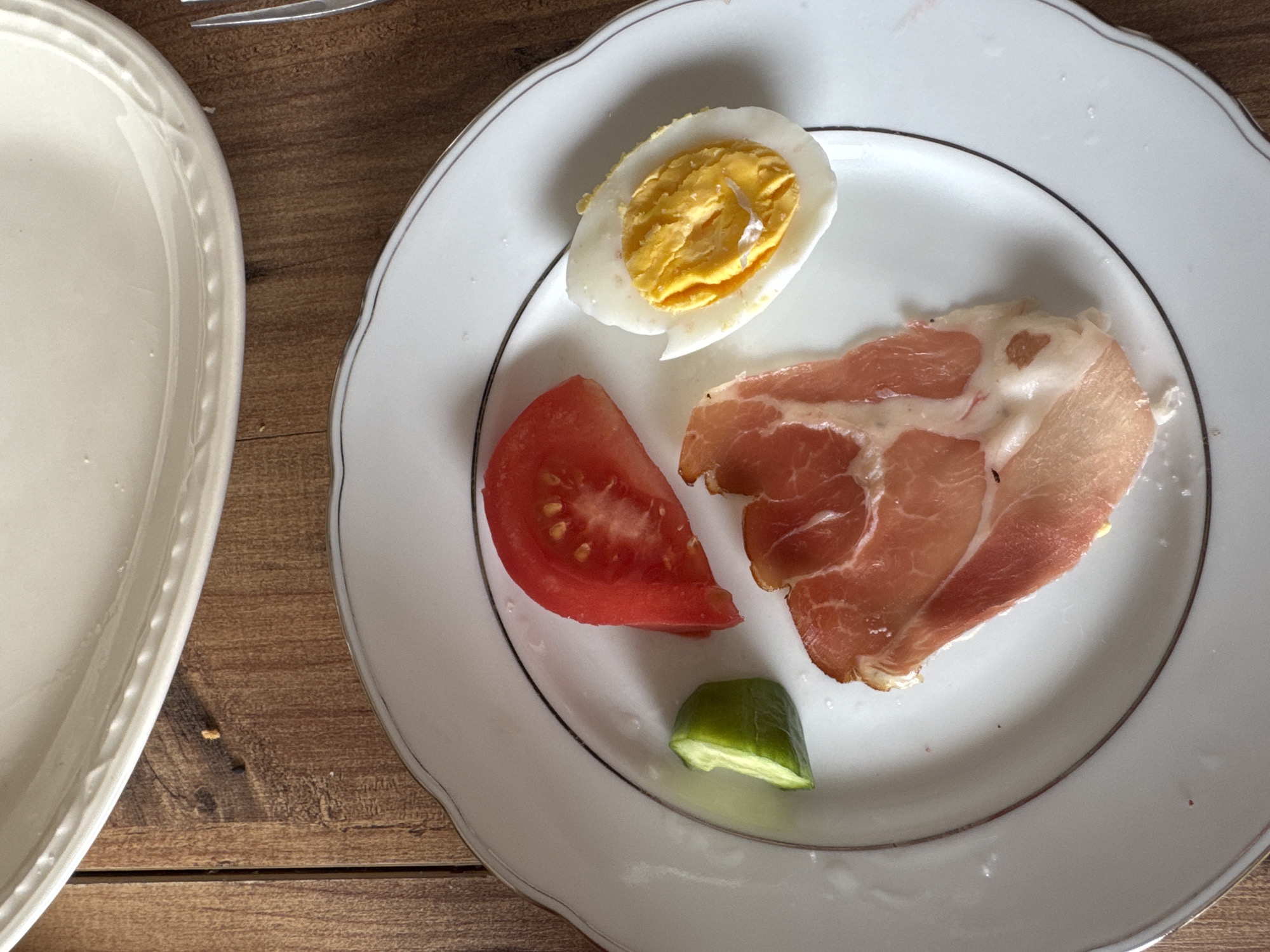 A white plate with a silver rim is on a wooden surface. The plate contains a halved hard-boiled egg, a slice of tomato, a slice of prosciutto, and a small piece of cucumber. Another white plate is partially visible on the left side of the image.
