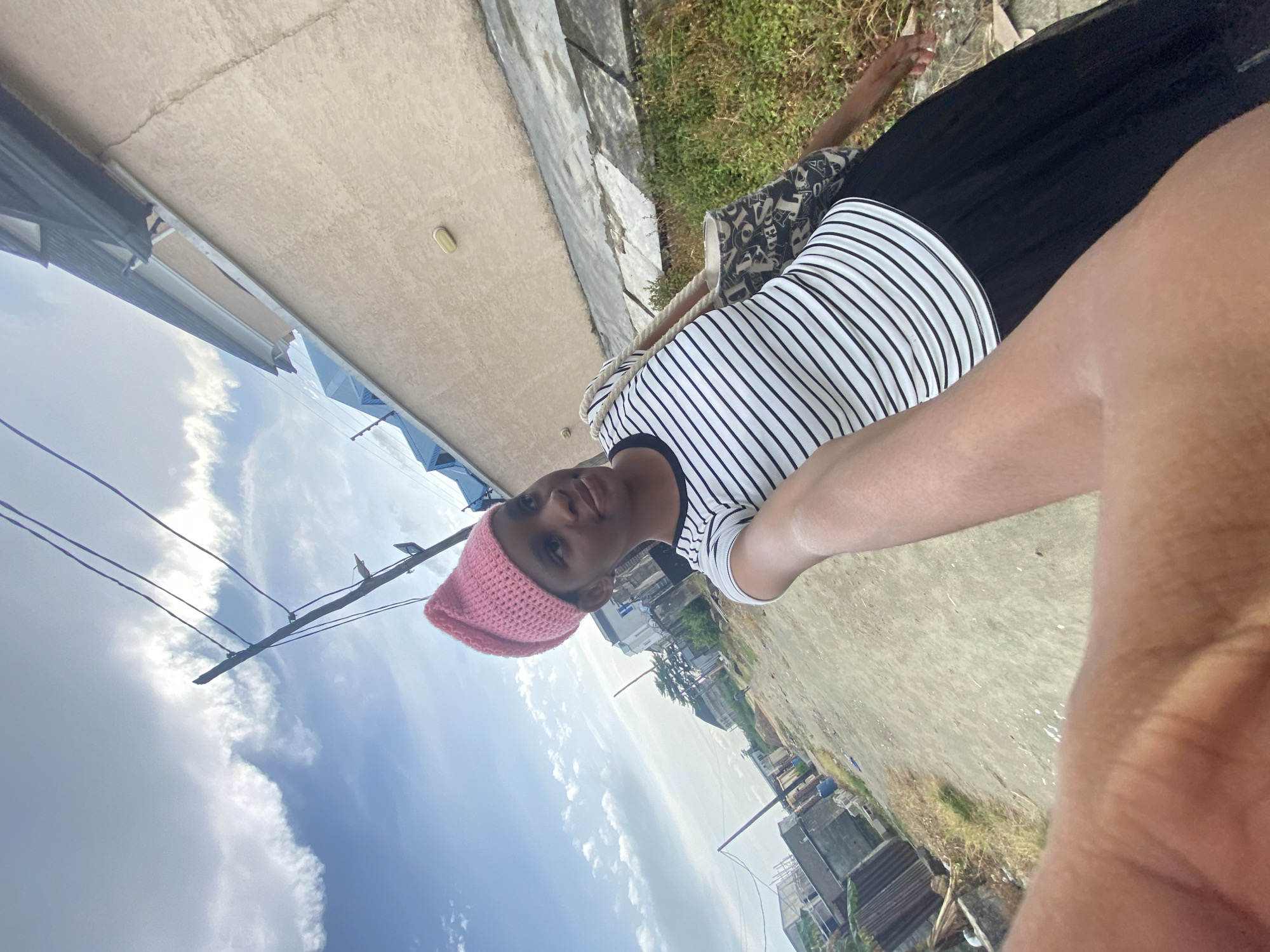 A person wearing a pink crocheted hat, a striped shirt, and a black skirt is visible in this outdoor shot. The person stands on a concrete surface in an urban environment. Buildings and greenery line the horizon under a sky filled with clouds and utility poles.