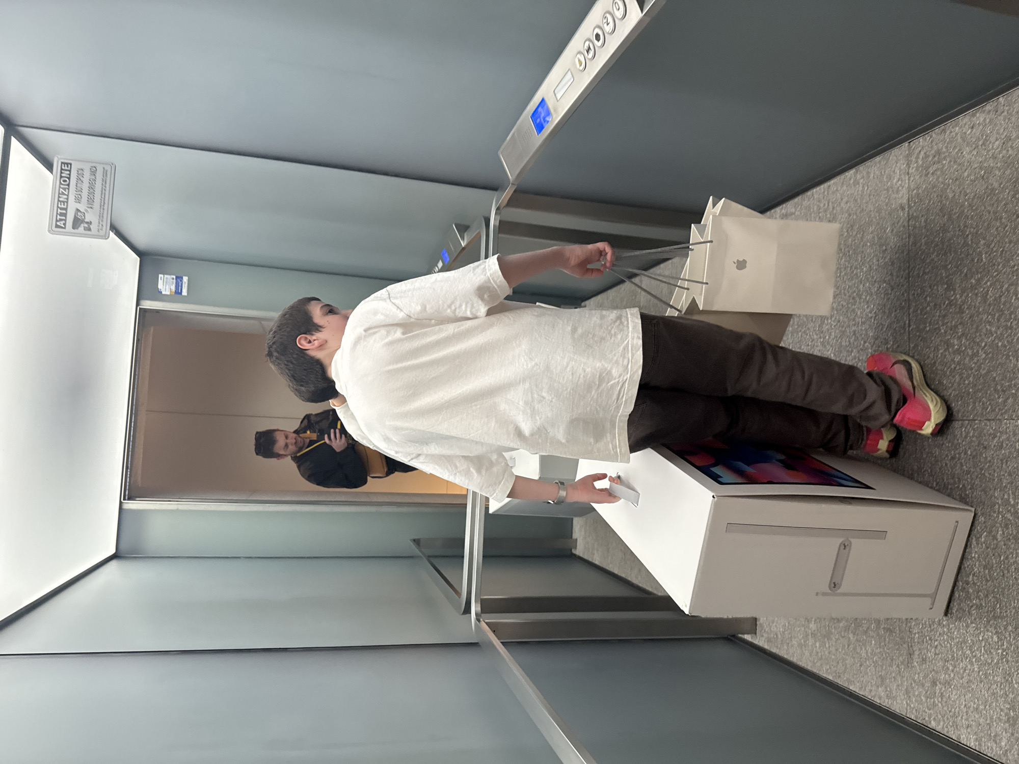 The image depicts a person exiting an elevator in what appears to be an Apple Store in Milan, Italy. The individual, dressed in a white long-sleeve shirt, brown pants, and pink sneakers, is carrying multiple white Apple shopping bags. They are stepping out of the elevator with a large white box displaying what appears to be an image of a computer screen on its top surface. Inside the elevator, a person in a black jacket with yellow accents is visible. The environment is characterized by sleek, modern architecture with light grey walls and metal accents, typical of an Apple Store. There is a sign with Italian text on the wall, and the elevator control panel is visible above. The floor is a speckled grey material.