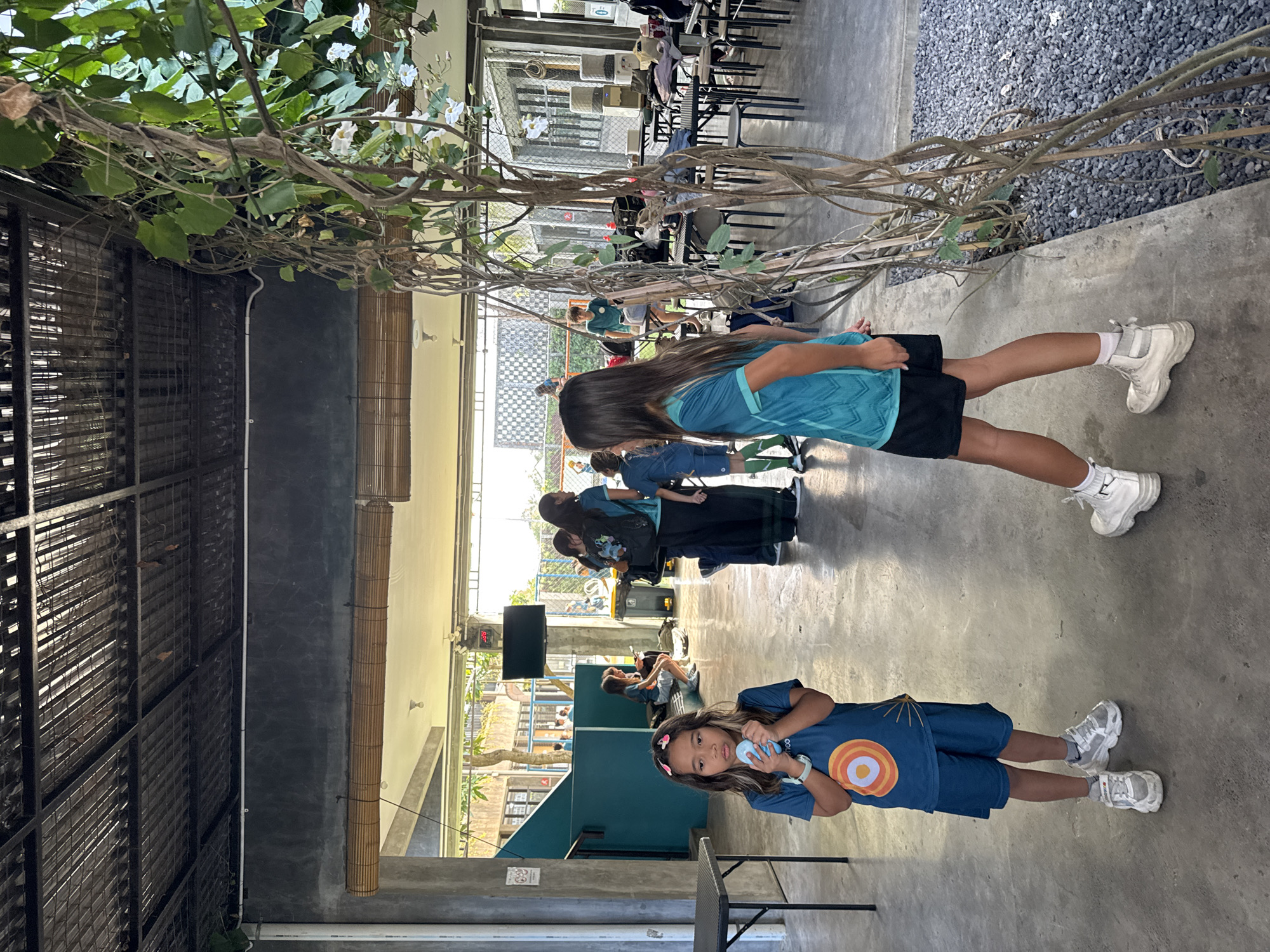 In North Kuta, Indonesia, an outdoor communal area with a grey concrete floor is depicted. A dark metal roof structure with exposed beams and grating covers the space, and bamboo blinds hang along a grey wall. Green vines with white flowers climb a wooden trellis in the foreground. Two young girls are the central focus. The girl on the left, wearing a teal t-shirt with an orange and white circular design and blue shorts, holds a light blue object to her mouth. She is also wearing grey and white sneakers and has her hair held back with pink clips. To the right, a second girl, facing away from the camera, wears a teal t-shirt with black shorts and white high-top sneakers. Several other people are visible in the background, some standing, some sitting at tables. Tables and chairs are arranged throughout the area, and a television screen is mounted on the wall.