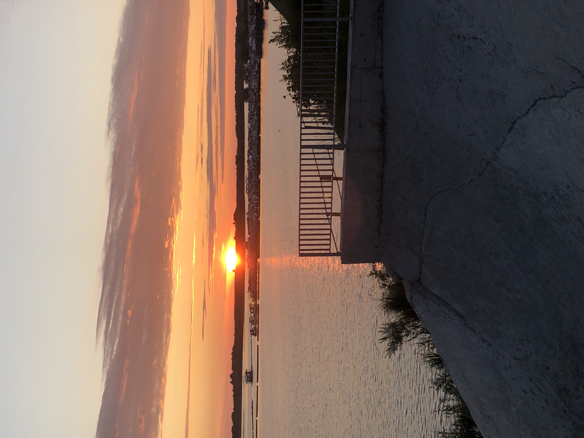 This image depicts a sunset over a body of water, likely a river or lake. The sky is painted with vibrant hues of orange, yellow, and pink, with scattered clouds. In the distance, a shoreline is visible with a line of trees and structures that suggest a populated area or a marina. Several small boats and floating structures are present on the water. In the foreground, there is a concrete embankment with a metal railing. The water reflects the colors of the sunset. The scene appears serene and tranquil. This location is not in Solikamsk, Russia; it is a generic waterfront scene.
