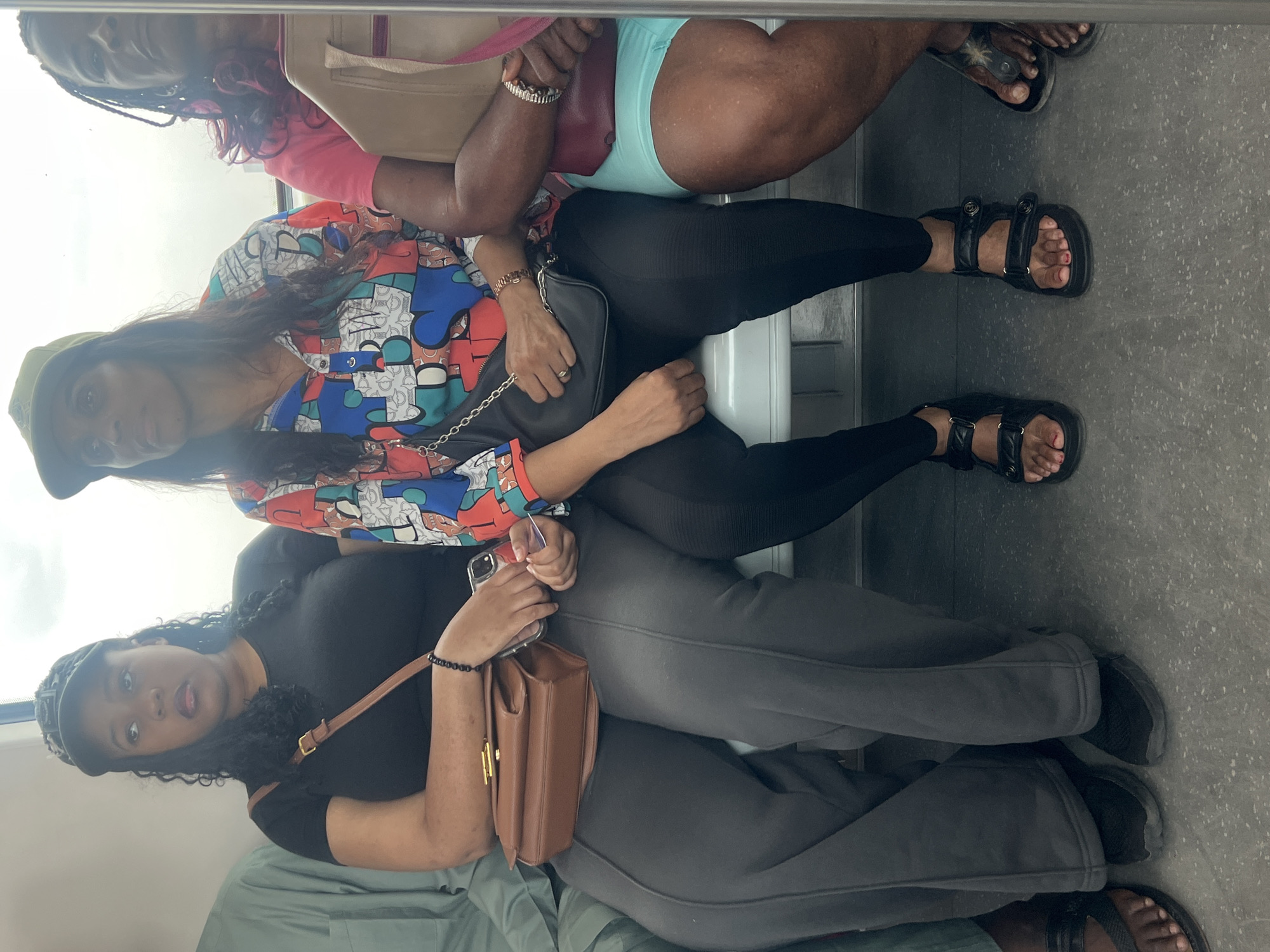 The image shows four individuals seated on a subway train. The location is speculated to be Aqani, Nigeria. The passengers occupy a shared bench. Starting from the top, one passenger wears a pink shirt and light blue shorts. Another wears a multi-colored shirt and black leggings. Another passenger is wearing a black shirt with gray sweatpants. Their attire suggests casual clothing.
Accessories include jewelry, handbags, and various footwear ranging from sandals to sneakers. They are engaging with smartphones. The environment is sparse, and only the subway seating and flooring are visible. The overall atmosphere is mundane.