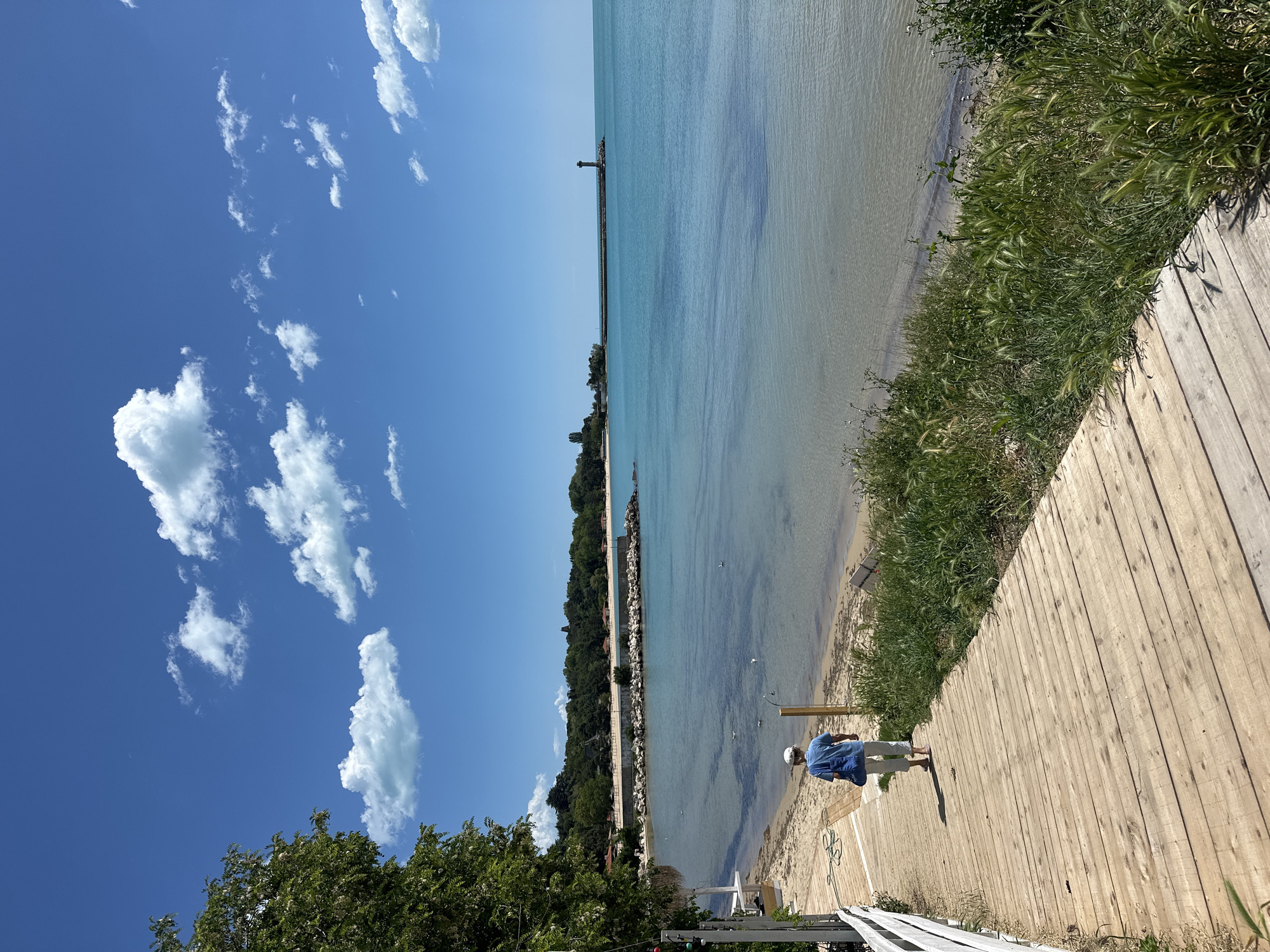 The image depicts a coastal scene in Varna, Bulgaria, featuring a boardwalk running parallel to the beach and sea. An individual wearing a blue shirt and light-colored pants is walking on the boardwalk. The sea is calm and clear, with a visible breakwater extending into the water. The sky is blue with scattered white clouds. Vegetation grows alongside the boardwalk, separating it from the beach. A building with a white fence is partially visible on the lower left corner of the image.