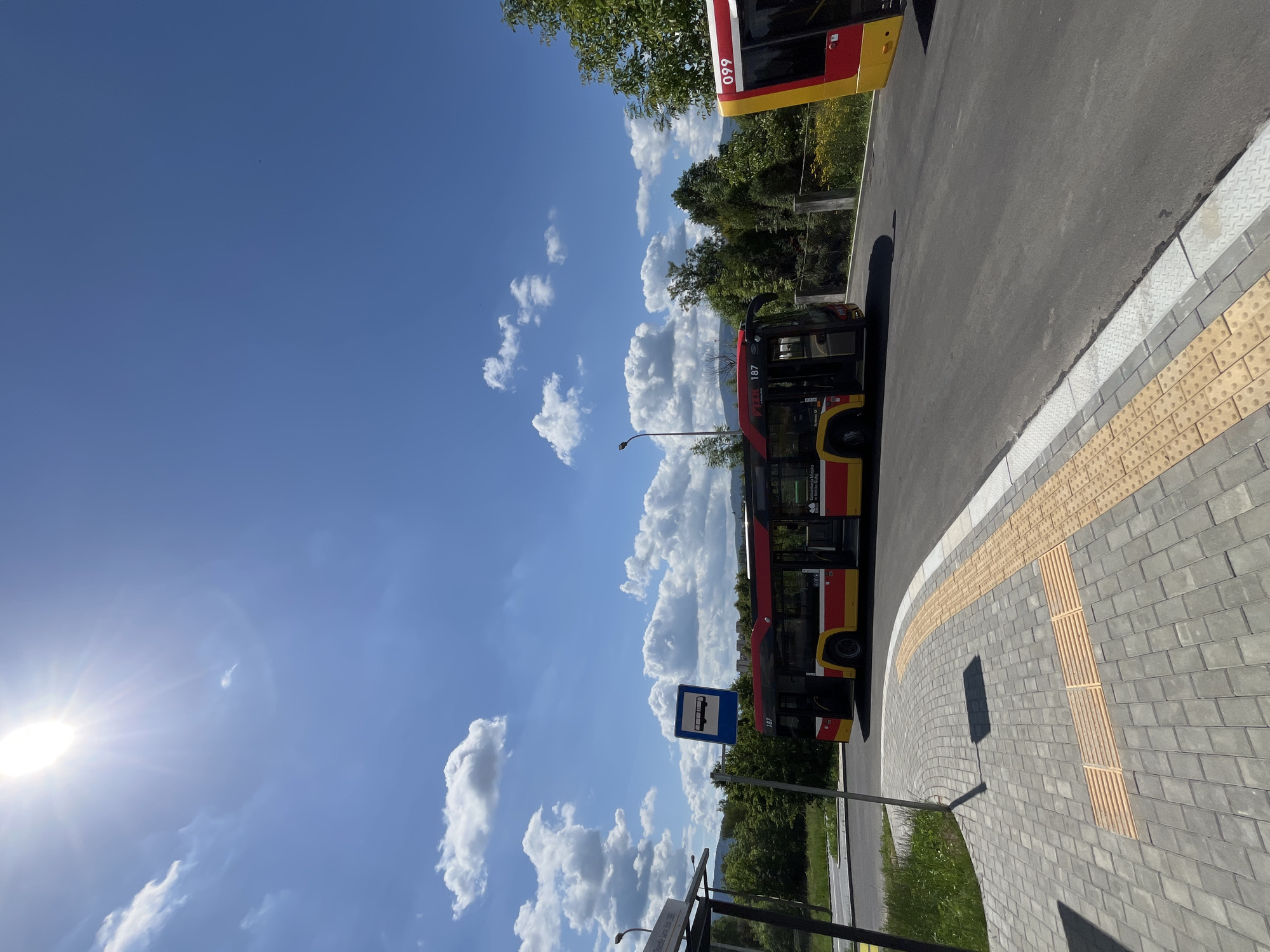 In the city of Unknown, Unknown, on a clear and sunny day with a bright sun and scattered clouds in a vibrant blue sky, a scene unfolds at a bus stop. A modern, articulated bus, painted in red and yellow, is parked with its doors open. The bus bears the number 187. To the right, the front portion of another similar bus, marked with 099, is visible. The bus stop features a sign with a bus icon. The ground in front of the bus stop is paved with gray interlocking stones, with a tactile paving system in yellow and gray for accessibility. A paved road with an asphalt surface leads away from the bus stop, bordered by a white line and a concrete curb. Lush green trees and foliage form a backdrop to the scene, with distant hills visible under the bright sky. The overall impression is of a functional public transport hub on a pleasant day.