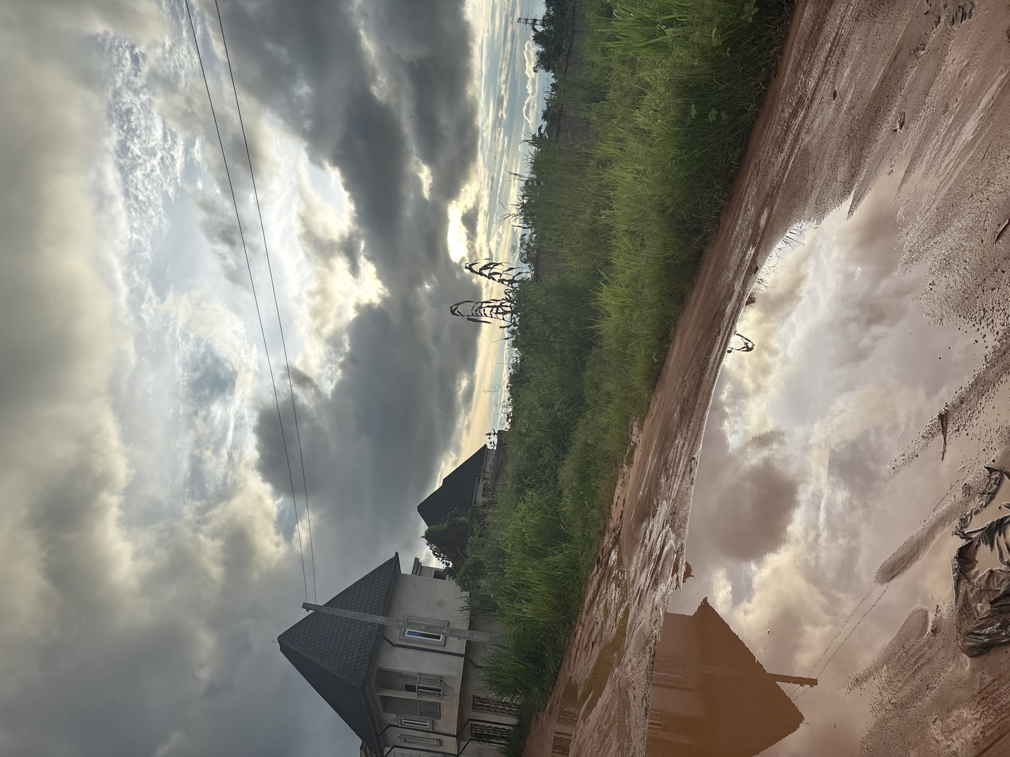 The image depicts an overcast sky with the sun peeking through the clouds, casting a diffused light on the landscape. In the foreground, a muddy, uneven dirt road leads away from the viewer. A large puddle of muddy water reflects the sky and the surrounding environment. To the left, a modern two-story building with a dark, pitched roof and balconies is visible, partially obscured by tall green grass. Behind this building, another similar structure with a dark roof is visible further in the distance, suggesting residential construction. The area is characterized by dense, tall green grass and vegetation, typical of a developing area in Obosi, Nigeria. The overall scene conveys a sense of rural or semi-urban development under a dramatic sky.