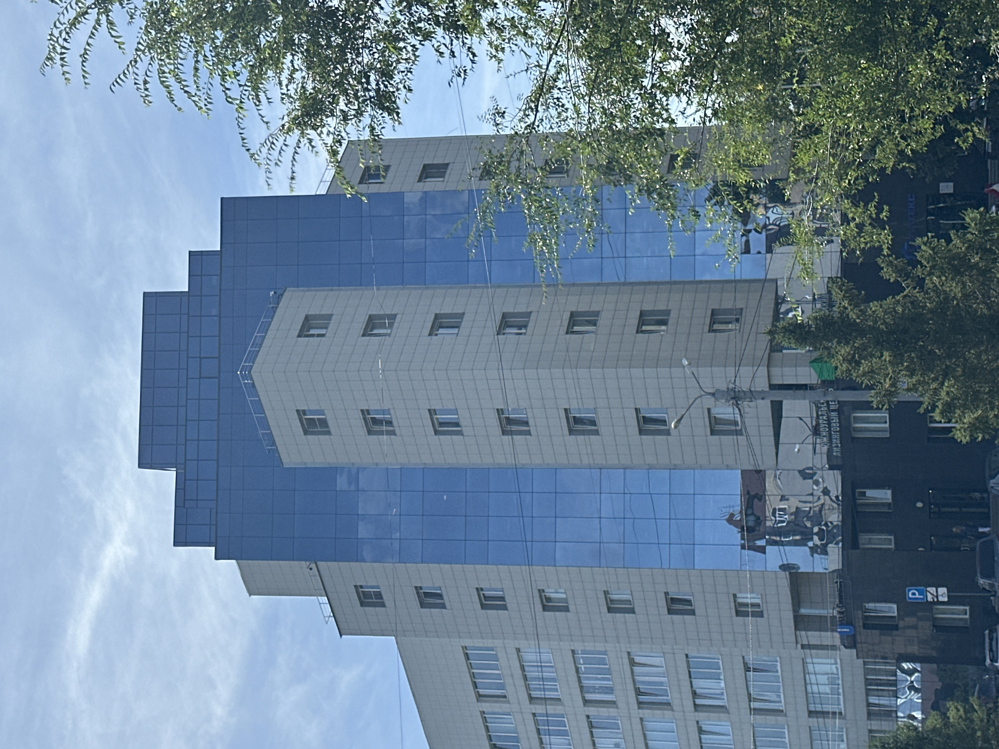 The image shows a modern, multi-story building with a stepped, tiered architectural design. The upper sections of the building are clad in reflective blue glass, while the lower sections are finished in a light-colored material with numerous windows. A darker, lower structure is visible at the base of the building, also featuring windows. Trees with green foliage partially obscure the view of the building. The sky above is a clear blue with wispy white clouds. A street sign with a "P" and a parking symbol is visible on the lower dark structure. Reflections of other buildings are visible on the blue glass facade. There are no people clearly visible in the scene. The overall environment appears to be an urban setting.
