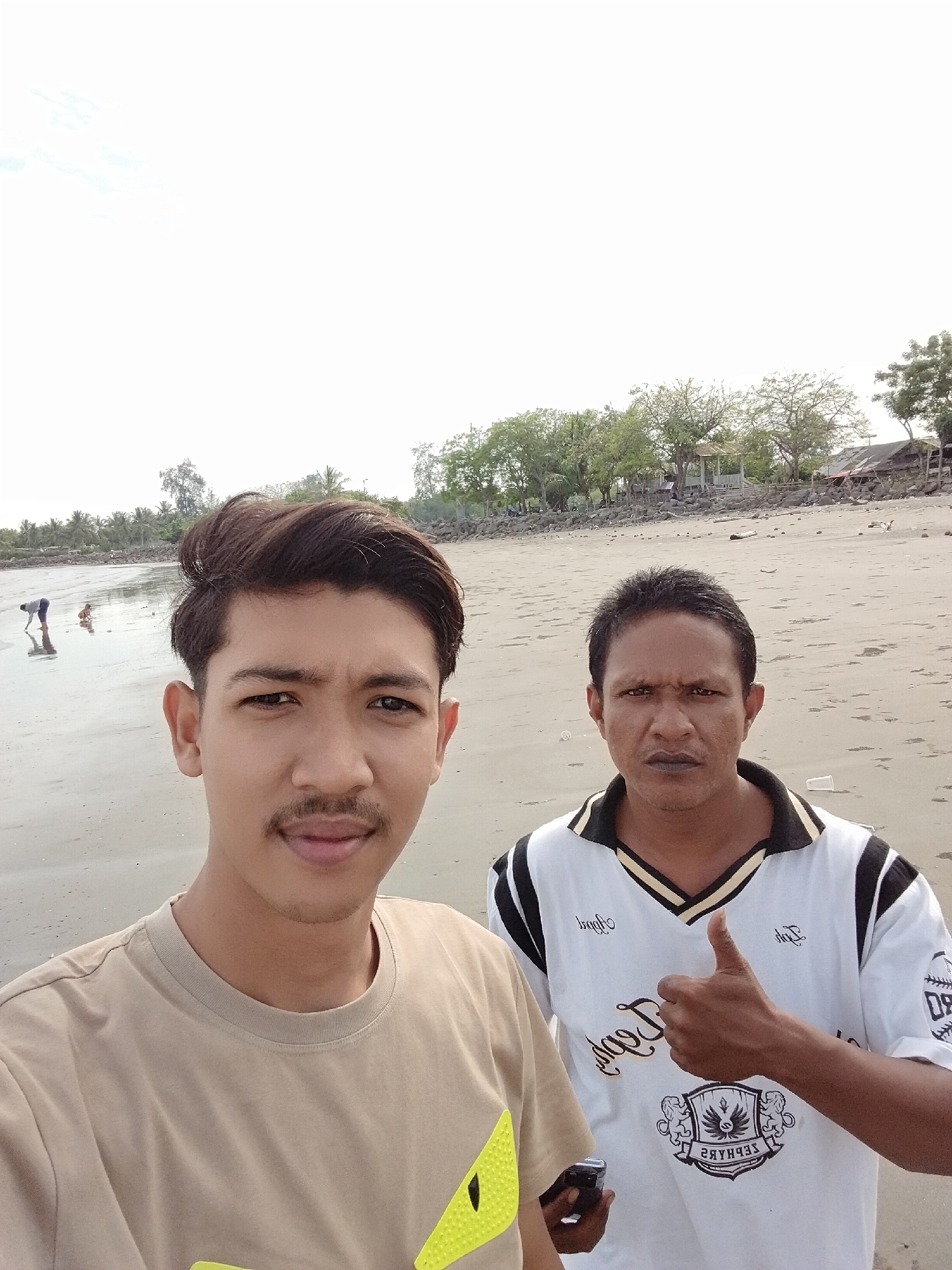 This selfie-style photograph features two men on a sandy beach in Lhokseumawe, Indonesia. The younger man is prominently positioned in the foreground on the left, looking directly at the camera with a slight smile. He has a short mustache and is wearing a light brown (or tan) round-neck t-shirt, part of a bright yellow pattern is visible on his lower right side.
Standing slightly behind and to the right is an older man, wearing a white polo shirt with black and gold striped trim on the collar and sleeves. He gives a thumbs-up gesture with his right hand and holds a black smartphone in his left hand. His expression is neutral to serious. Due to the selfie orientation, the text on his shirt is mirrored: a prominent crest on the chest features the words "JEJAK" and "ZEPHYRS" (reversed), with additional reversed text such as "Jejak" and "dirq" scattered across the shirt.
The background reveals a wide, light-colored sandy beach, with wet areas reflecting the sky, suggesting low tide. In the distance, near the water's edge, two smaller figures, an adult bending over and a child, are visible. The shoreline beyond is lined with various trees, including some tropical palms and leafy trees, interspersed with simpler coastal structures and a small gazebo-like building. Rocks are also present along the distant shoreline. The sky is bright and appears to be overcast or hazy, diffusing the sunlight. A few plastic cups and other small pieces of debris are scattered on the sand. The overall impression is a daytime scene on a coastal setting.