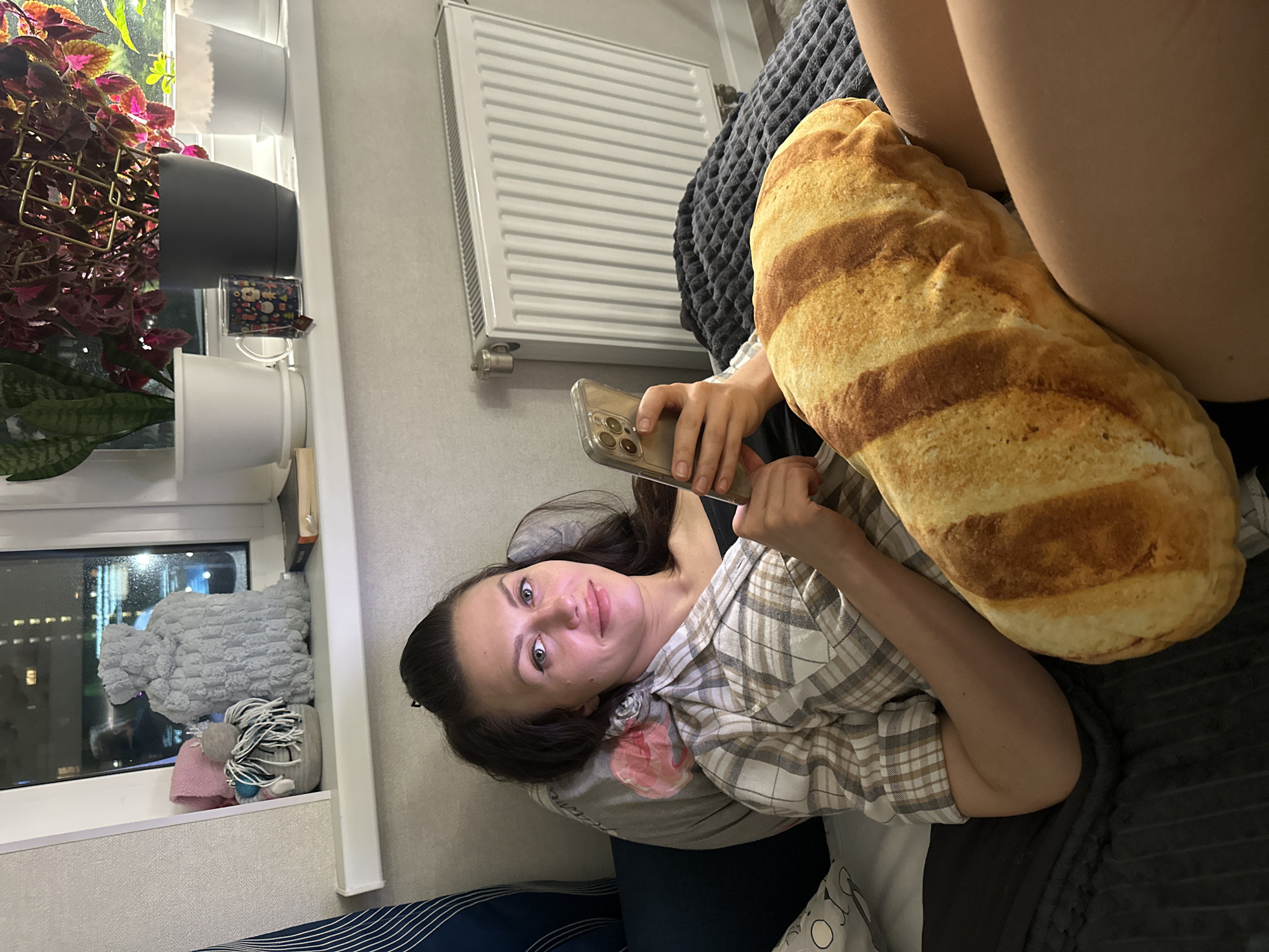 A young woman is captured in a relaxed indoor setting, likely a home in Murino, Russia. She is lying down, positioned to look towards the camera with a gentle, content smile on her face. Her dark hair frames her face, and she has blue-grey eyes. She wears a light-colored plaid shirt over a dark top and is holding a smartphone with a clear case in her hands.
Beside her, on a dark, textured blanket, rests a large, realistic baguette-shaped pillow. In the background, a white radiator is mounted on the wall. To the left, a window reveals a dark outdoor scene with faint distant lights, indicating that it is nighttime or evening.
The windowsill is adorned with several houseplants, including one with vibrant red and green foliage (possibly a coleus plant) and another with green, pointed leaves resembling a snake plant. A dark mug with small, colorful patterns is also visible on the windowsill, alongside a light grey, deeply textured throw or pillow and what appears to be the spine of a book. The overall atmosphere is cozy and domestic. No discernible text is visible on any objects.