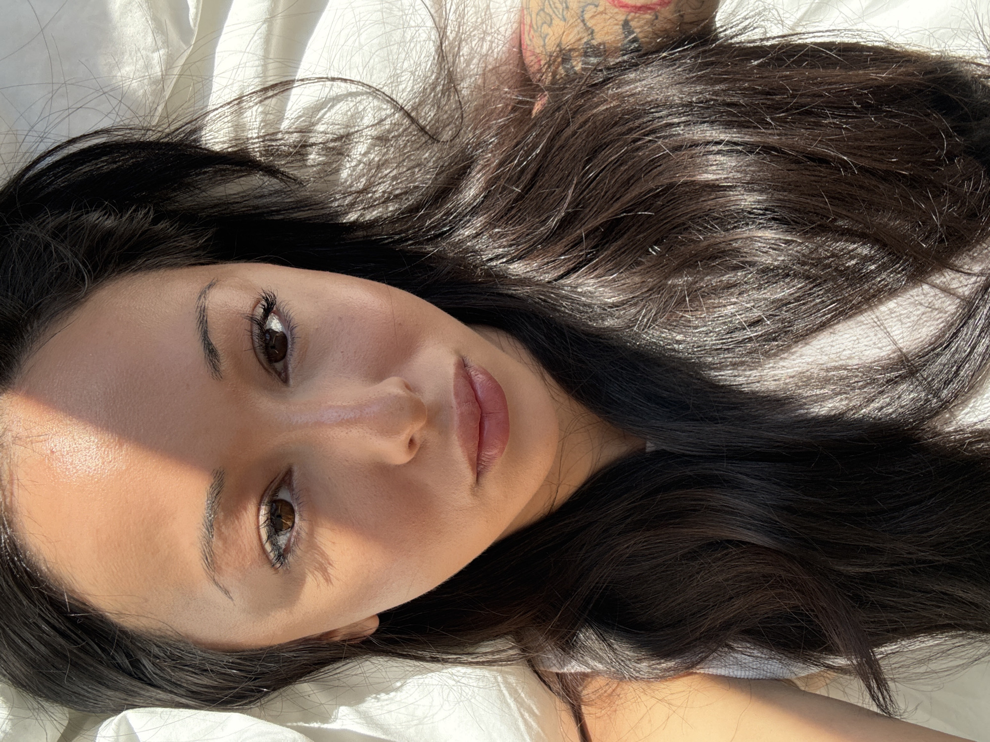 A portrait features a young woman lying down, looking directly upwards at the camera. Her head is positioned on a white, crumpled fabric, likely a bedsheet or pillow. She has long, dark brown hair, which is spread out around her head, and striking dark brown eyes. Her skin appears smooth and well-lit, with a natural sheen visible on her forehead and cheekbones. Her lips are slightly parted, revealing a soft, natural pink color.
Bright, natural sunlight streams from an unseen window, creating distinct, sharp shadows across her face and highlighting parts of her hair and skin. This strong illumination suggests it is daytime, possibly morning or late afternoon. Her expression is calm and serene.
An intricate tattoo, featuring patterns with hints of red or orange coloration, is visible on her upper right arm or shoulder, partially obscured by her hair. The immediate background consists solely of the white fabric. There are no explicit visual cues within the image to confirm the specified location of London, United Kingdom; the setting appears to be an indoor domestic environment.