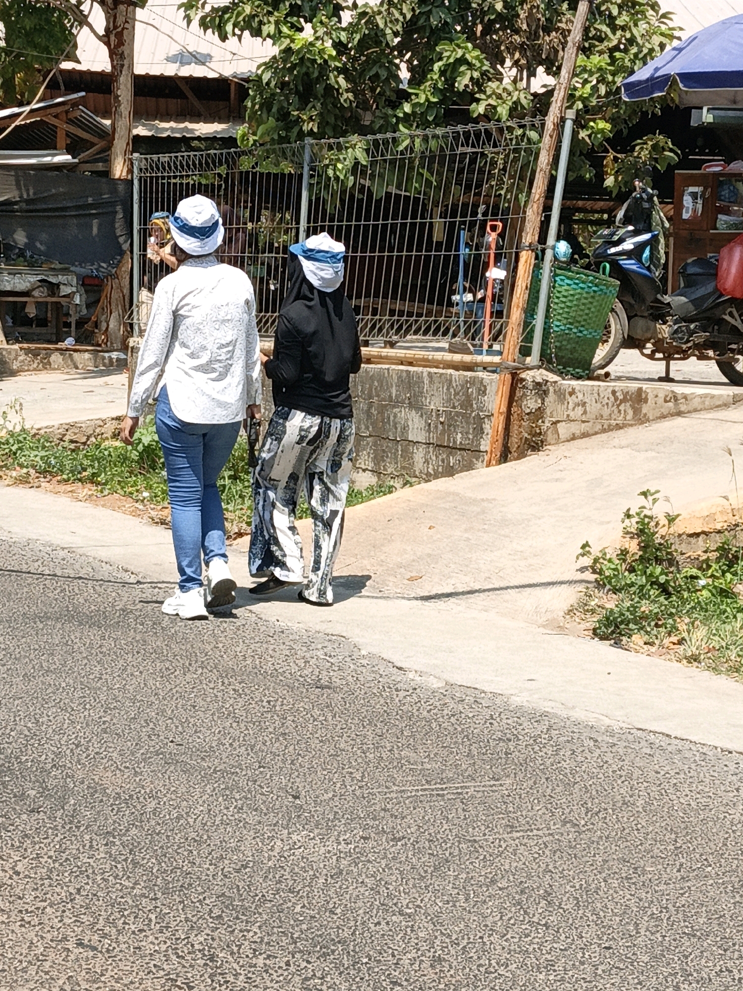 Two individuals, viewed from the rear, walk along a paved street in Kuwasen, Indonesia, on a sunny day. The individual on the left wears a white long-sleeved shirt with a subtle patterned print and blue jeans, paired with white sneakers. The person on the right is dressed in a black long-sleeved top and patterned pants with black and white designs, also wearing dark shoes. Both individuals have white head coverings with blue trim. They are walking away from the camera on a sidewalk that runs adjacent to the street. In the background, a metal fence encloses an area with vegetation and structures. A motorcycle is parked on the right side of the image near a blue umbrella and a wooden structure. The environment appears to be a semi-urban or rural setting, characterized by paved surfaces, a fence, greenery, and buildings.