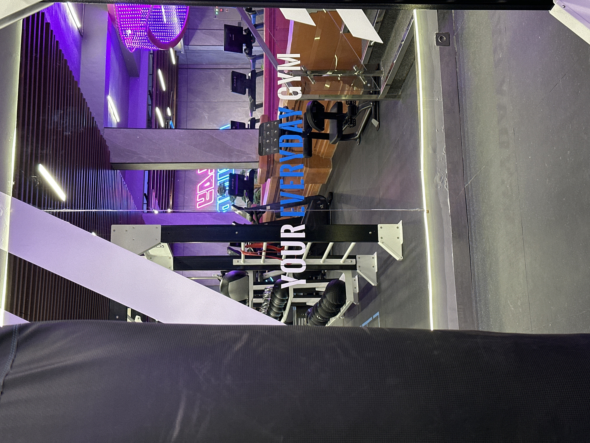 The image depicts the interior of Everyday Gym, a fitness facility located in North Kuta, Indonesia. The gym features modern decor with purple and white lighting elements and wooden slat accents. There are exercise bikes visible in the background and foreground. Signage with the phrases "EVERYDAY GYM" and "YOUR" are prominently displayed. The lower portion of the image is dominated by a dark, textured surface, possibly part of the equipment or a partition within the gym. There are no individuals directly visible in the frame.