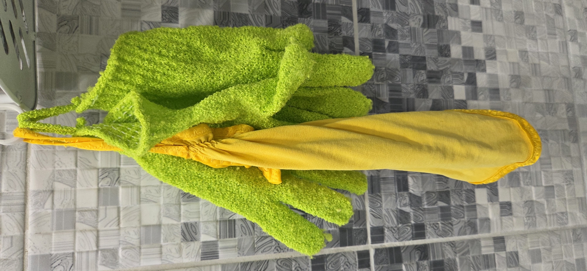 Two bright lime-green, textured exfoliating gloves and a folded, bright yellow cloth hang from a white plastic hook on a tiled wall. Each glove features a nappy, looped surface and a small loop at its cuff for suspension. The yellow cloth is cinched and positioned between the two gloves, sharing the same hanging point. The background wall is composed of small, square ceramic tiles, alternating between lighter and darker gray shades, each featuring a subtle, abstract swirling pattern. The tiles are arranged in a grid with visible light-colored grout lines. The scene is evenly lit, indicating an indoor environment. The location is the city of Lipetsk, Russia.