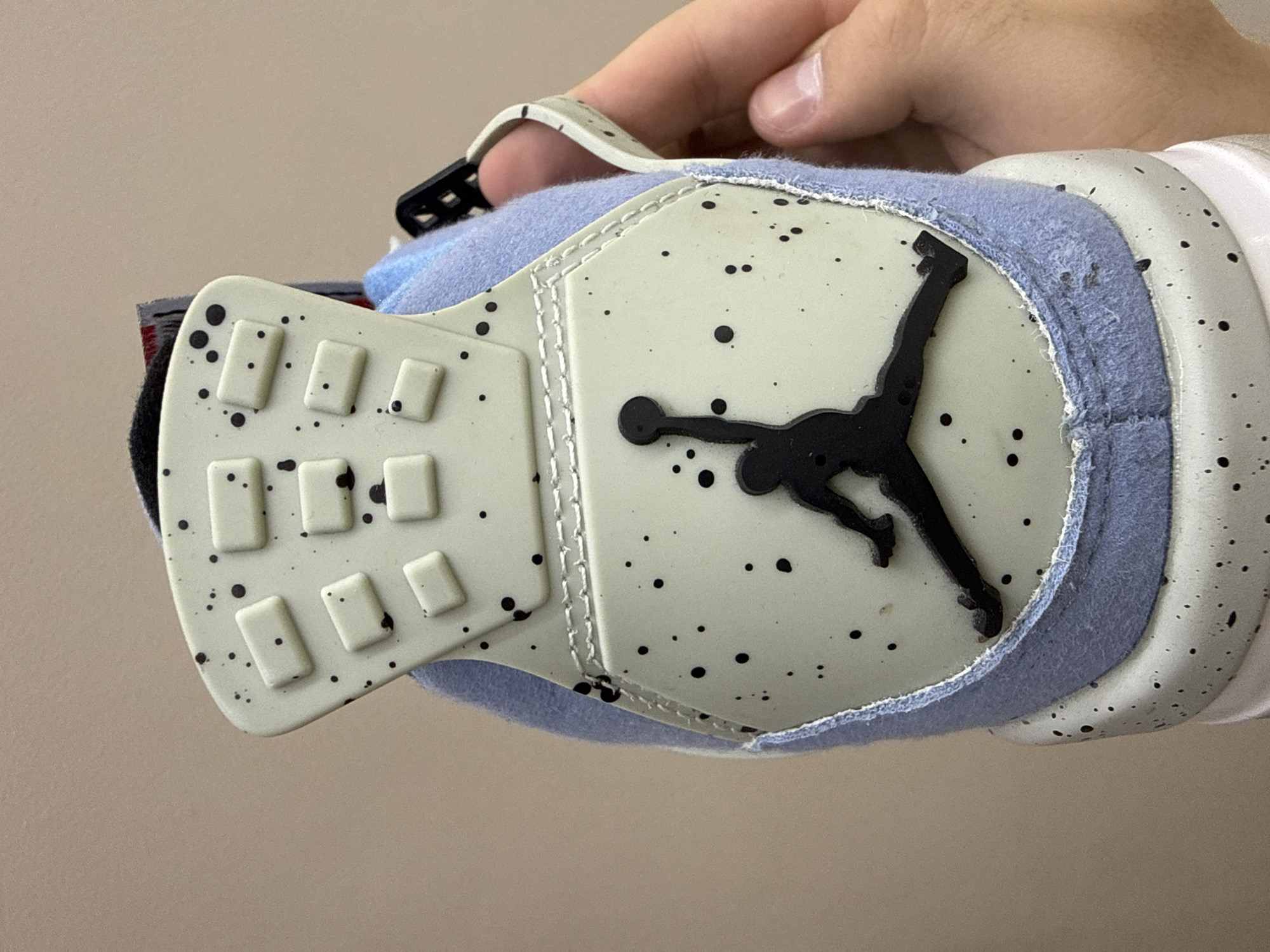 The image shows a close-up of a hand holding a shoe. The shoe appears to be a sneaker, with a light-colored base speckled with black dots. A black Jumpman logo is prominently displayed on the side of the shoe. The shoe also features a light blue fuzzy material around the top edge and a white strap with a black buckle. The background is a plain, light beige color, suggesting an indoor setting with neutral lighting. There are no discernible location cues within the image itself, and no text is visible. The focus is entirely on the details of the shoe and the hand holding it. The image is a product shot, likely intended to showcase the design and features of the footwear.