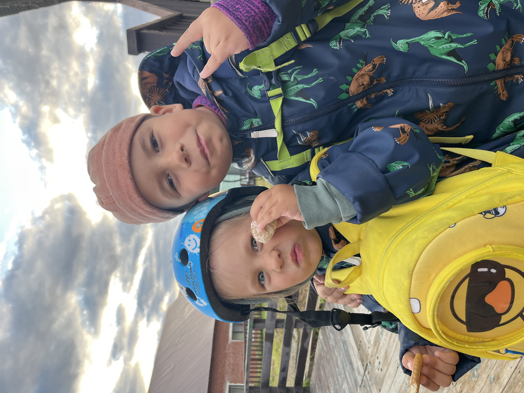 Two children, a boy and a girl, are standing close together in front of a wooden structure and a brick building under a cloudy sky. The girl, on the left, is wearing a blue helmet with a smiley face design and a yellow backpack. She is holding a small biscuit up to her eye. The boy, on the right, is wearing a reddish beanie and a dark blue jacket with a dinosaur print. He is making a rock-and-roll hand gesture with his right hand and has his left arm around the girl. The environment appears to be outdoors, possibly in a park or recreational area, given the wooden walkway and fence. There is no specific visual information in the image that points to the city of Chaikovsky, Russia.