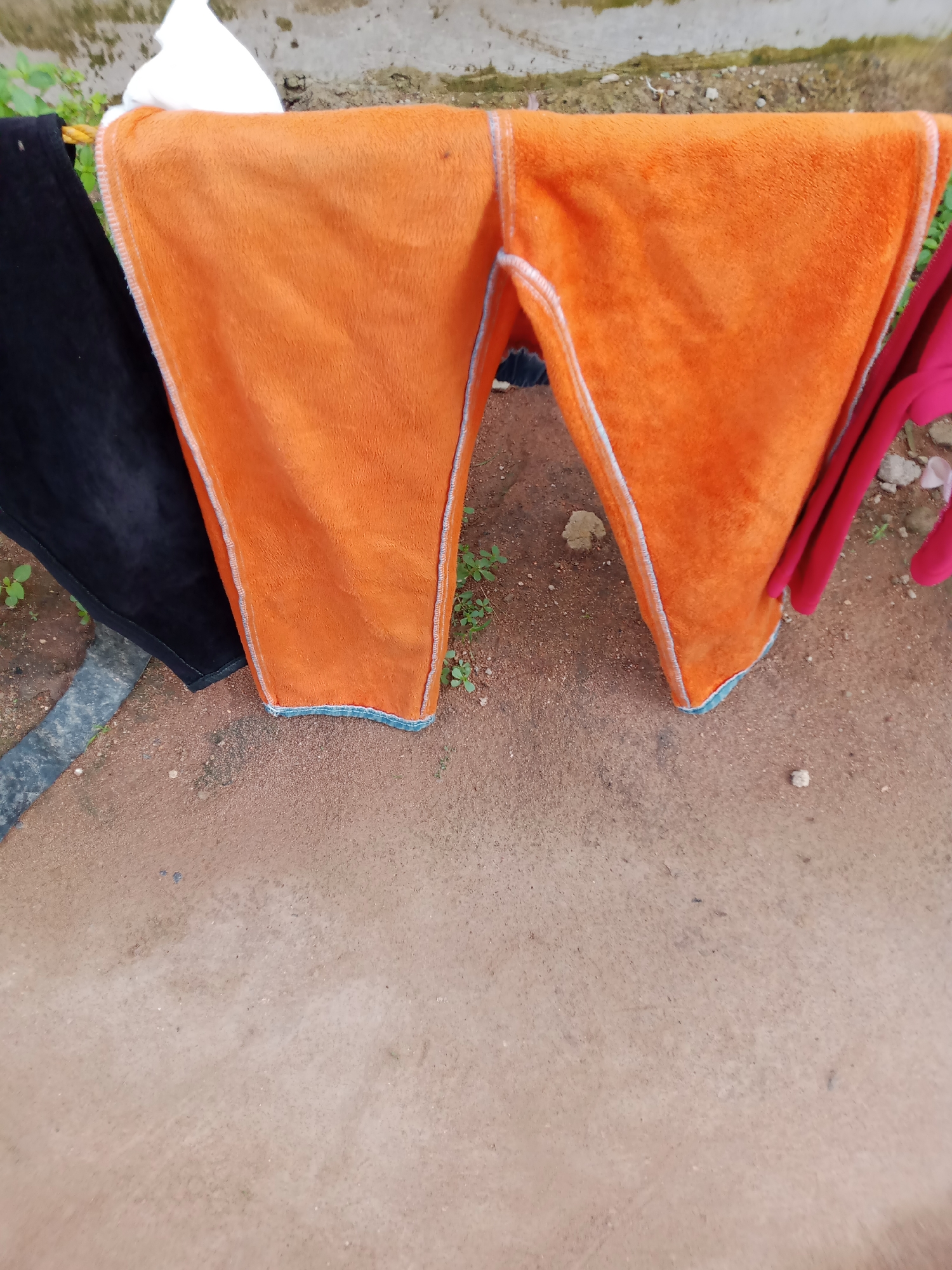 In Jauro Yinu, Nigeria, an outdoor clothesline displays various items of clothing. On the left, a pair of black trousers hangs. Adjacent to the black trousers is a pair of bright orange pants, distinguished by a light blue and white trim along the seams. To the right of the orange pants, a piece of dark red fabric is visible, possibly another garment. The clothing is suspended from a rope, with a mottled concrete wall and sparse greenery forming the background. The ground beneath is a reddish-brown dirt surface, scattered with small stones and pebbles. The overall scene suggests laundry being air-dried under natural conditions.