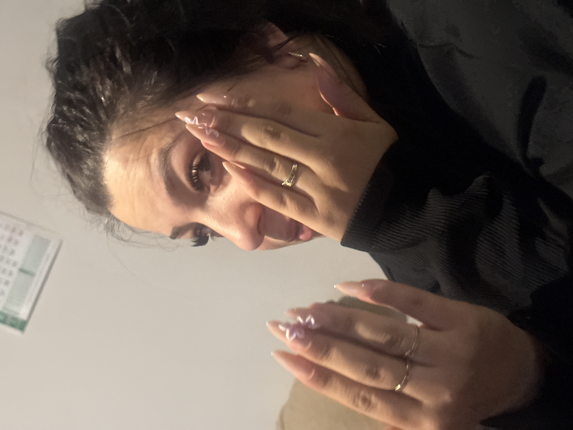 A young woman with dark hair, styled upwards and back from her forehead, is partially obscuring her face with her hands. She is wearing a dark-colored, ribbed-knit sweater or jacket. Her nails are long, manicured with a light pink base, and have small, white flower designs. She is wearing a ring on one of her fingers. A calendar is visible in the background, out of focus, partially obscured by a corner of the room, and appears to be mounted on a light-colored wall. The scene appears to be indoors with low lighting. No details indicate the location is in Chubra, Bulgaria.